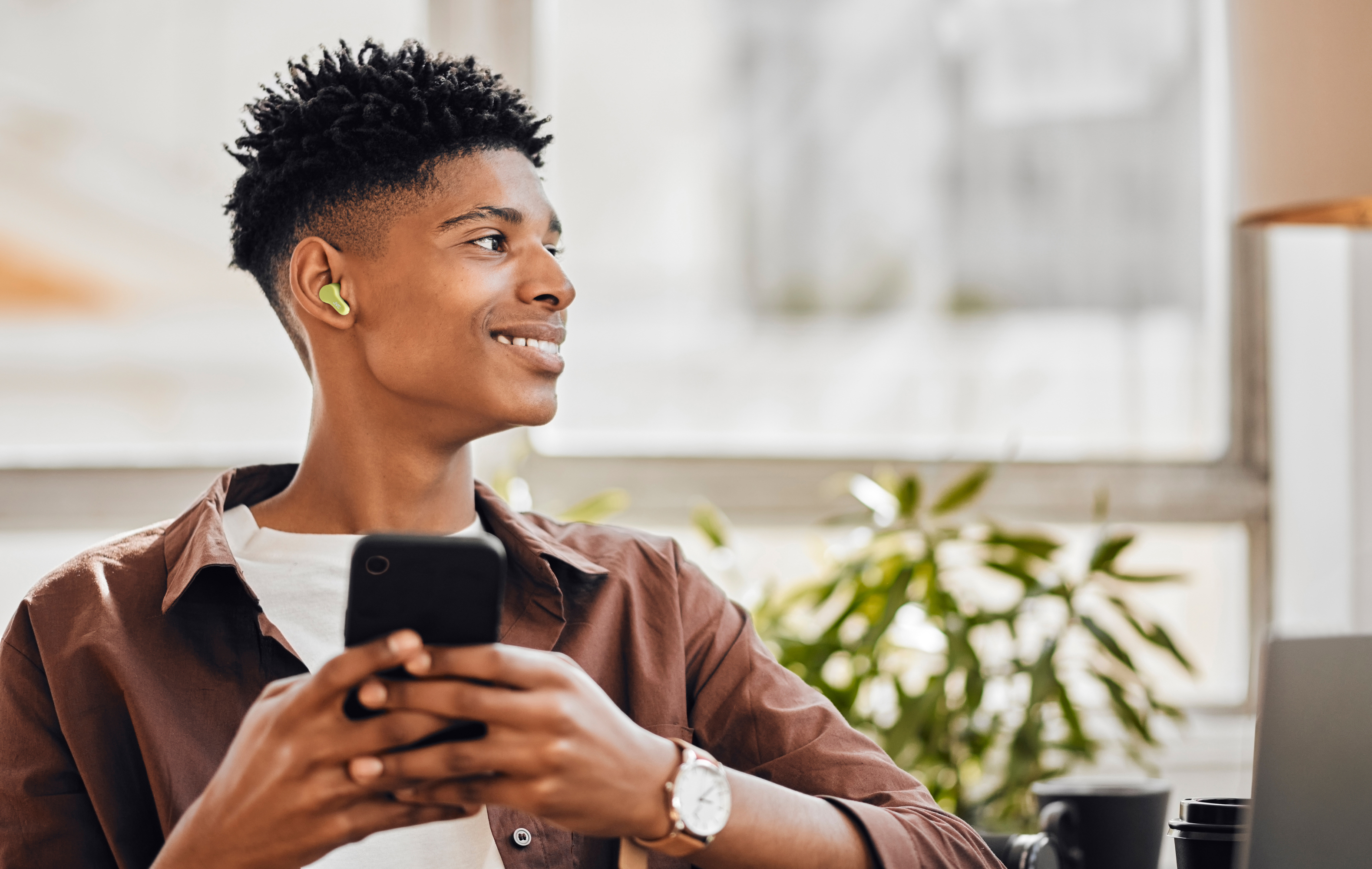 Un hombre negro sonriente con auriculares y un teléfono, con una camisa marrón. Mira a la derecha, con fondo borroso.