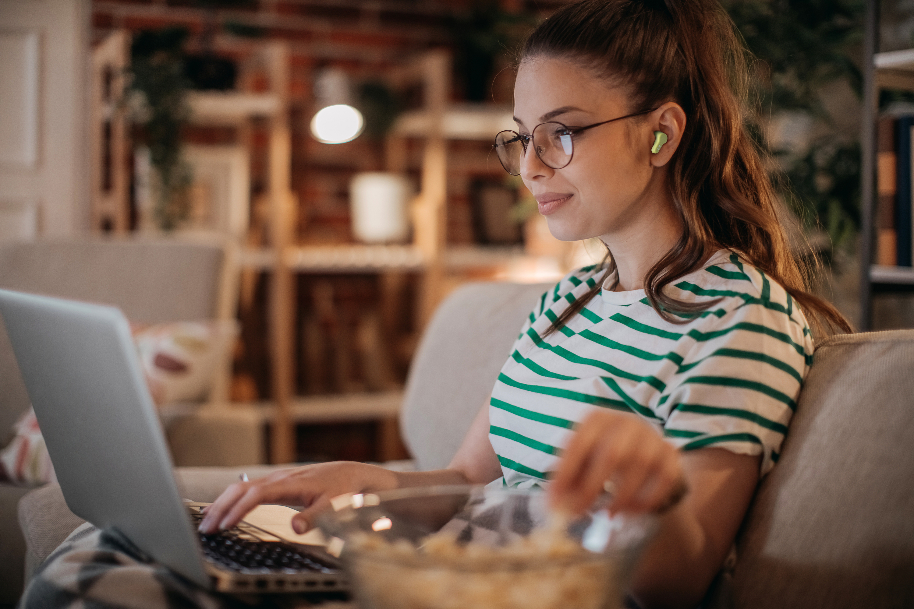 Una mujer con gafas, auriculares verdes y una camisa a rayas usa una laptop en un sofá. Un tazón de palomitas.