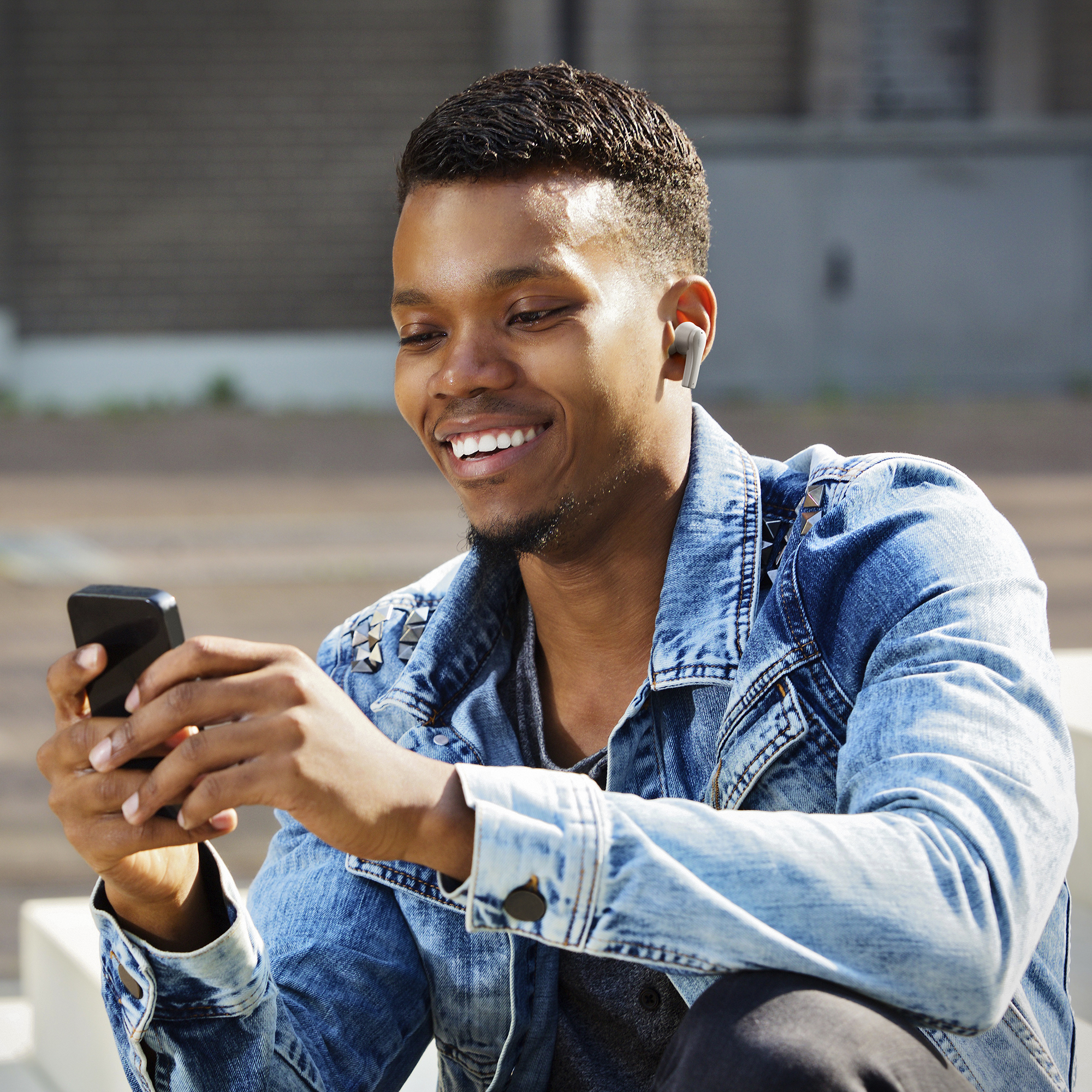 Lächelnder Schwarzer Mann mit Ohrhörern, hält ein Telefon, trägt eine Jeansjacke. Urbaner Hintergrund.