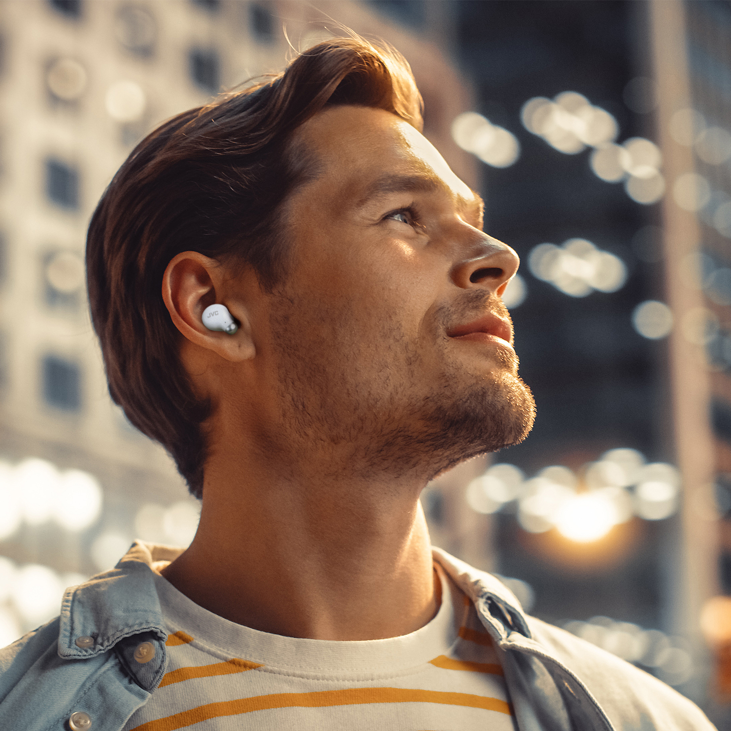 Un uomo con un auricolare guarda in alto, con le luci della città sullo sfondo. Indossa una maglietta a righe e una giacca di jeans.