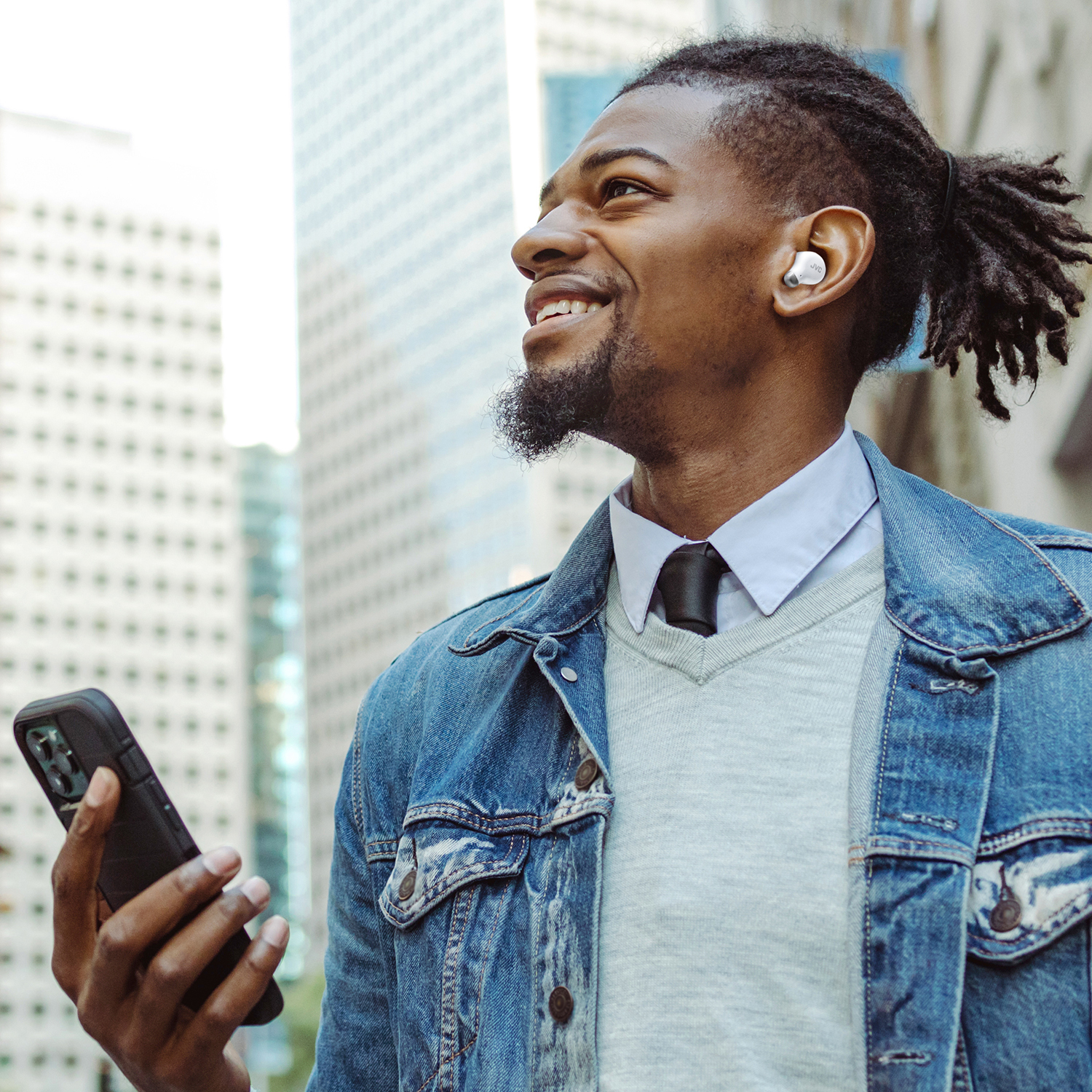 Un uomo nero sorridente con una giacca di jeans tiene un telefono, indossa un auricolare, con grattacieli sullo sfondo.