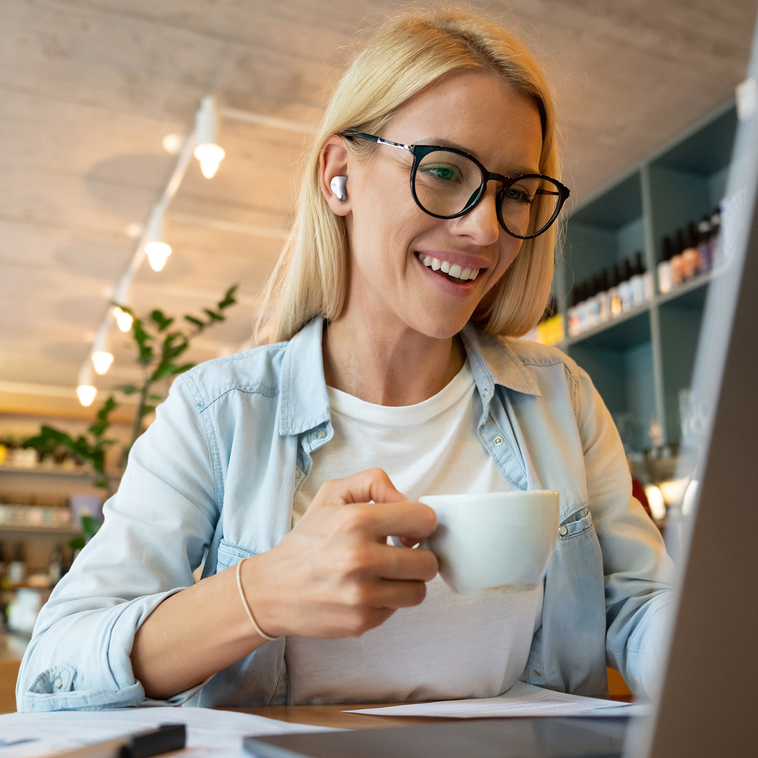 Donna bionda con occhiali sorride mentre guarda un laptop. Tiene una tazza di caffè. Lo sfondo include l'arredamento del bar.