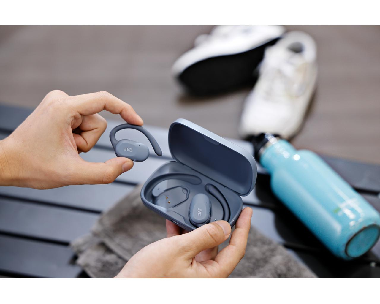 Ręce trzymają szare słuchawki JVC i etui. Buty sportowe i butelka z wodą w tle. Ręcznik na czarnym stole.