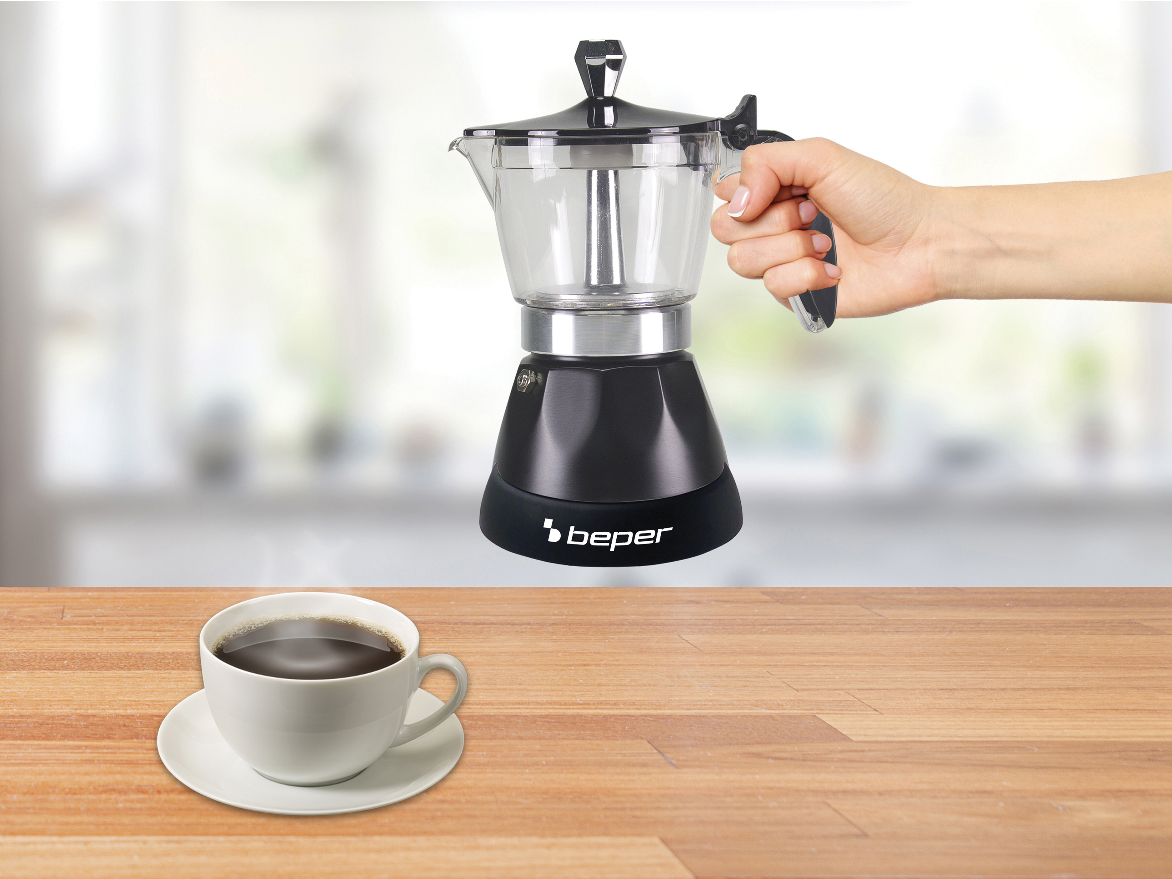 Una mano tiene una caffettiera nera e trasparente. Una tazza di caffè è su un tavolo di legno. Lo sfondo è una cucina sfocata.