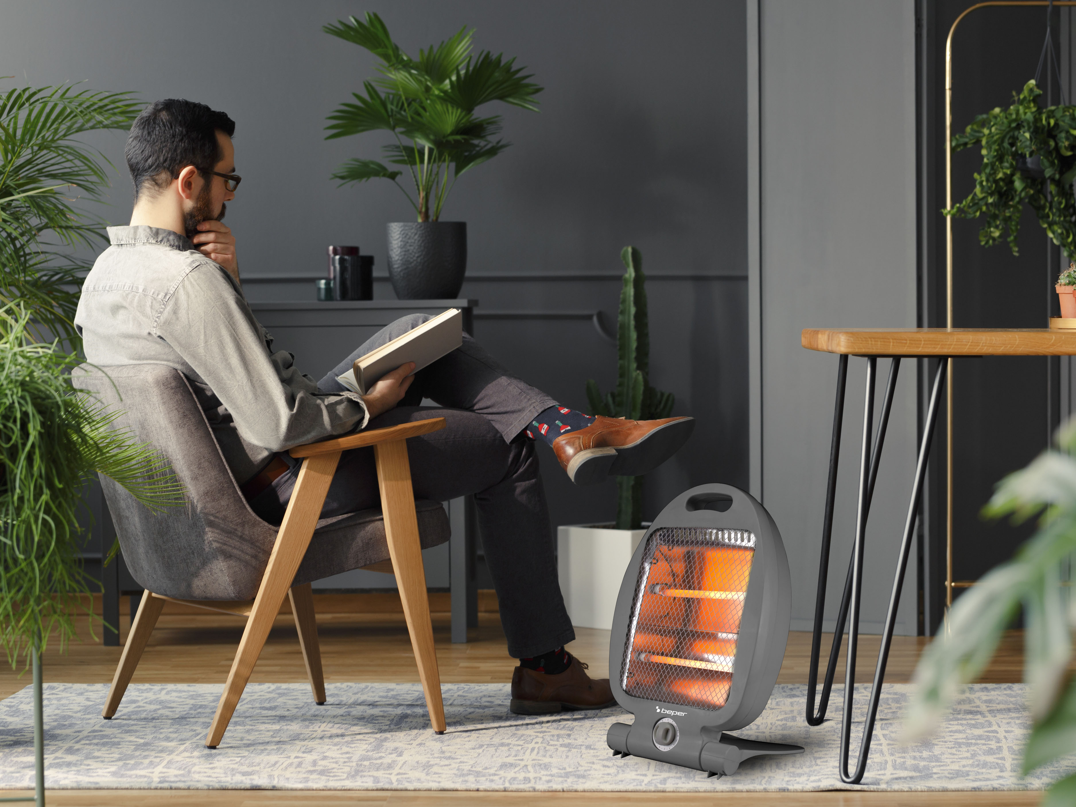 Un uomo su una sedia grigia legge un libro. Un termoventilatore è di fronte. Ci sono piante intorno.