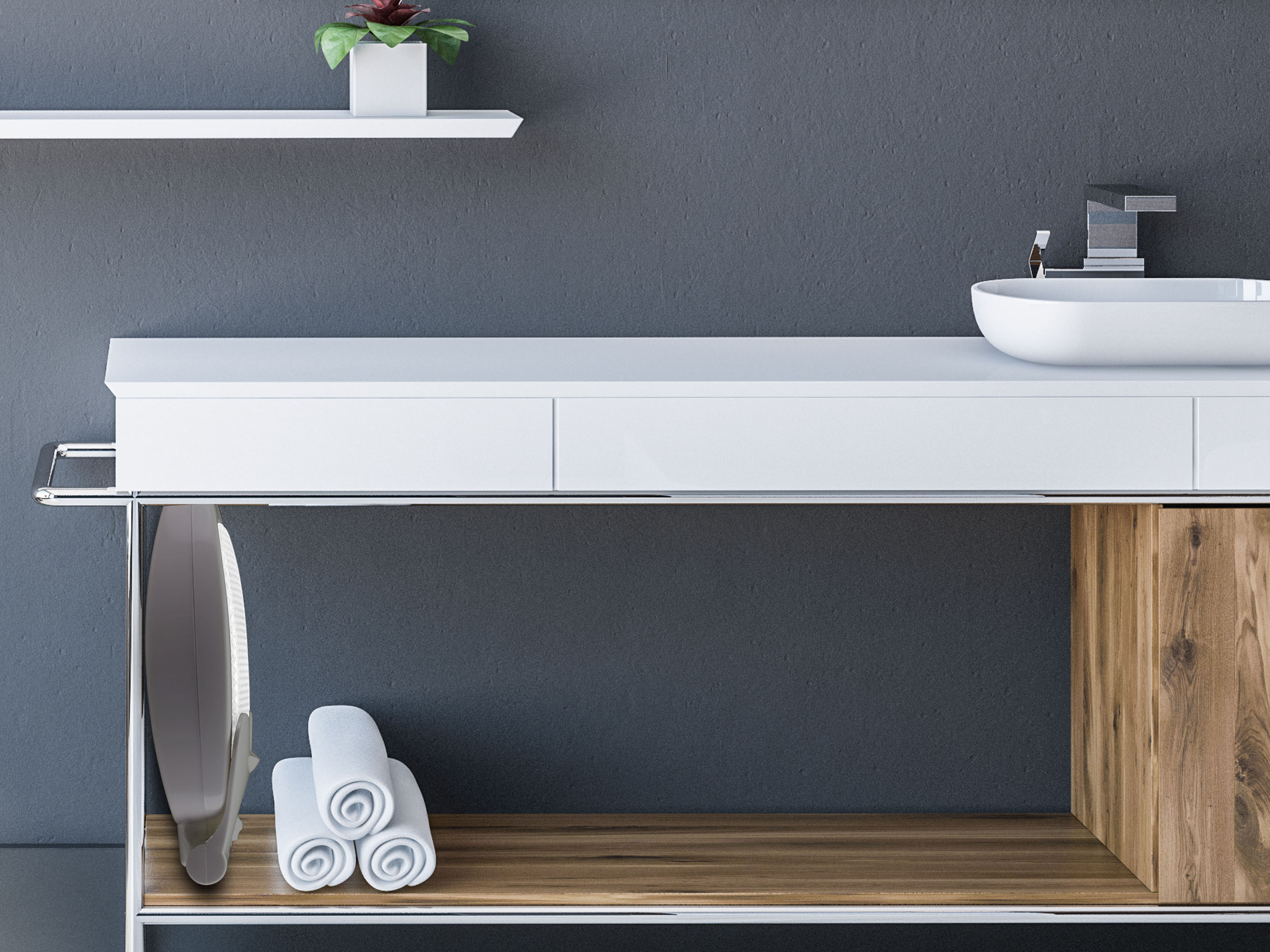 Bagno con lavandino bianco, mobile in legno, asciugamani arrotolati e parete grigia. Una pianta è su una mensola.