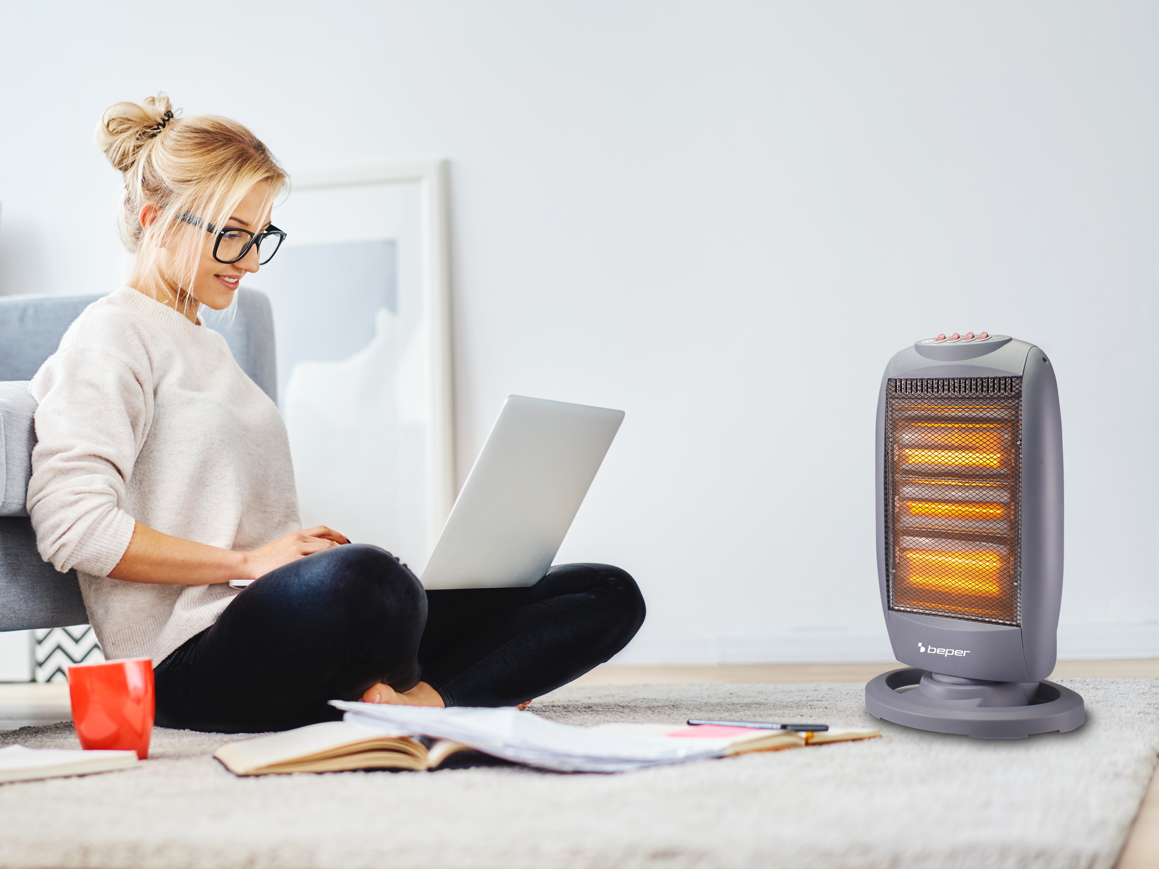 Donna con laptop seduta vicino a un termosifone. Una tazza rossa e un libro in primo piano. Indossa occhiali.