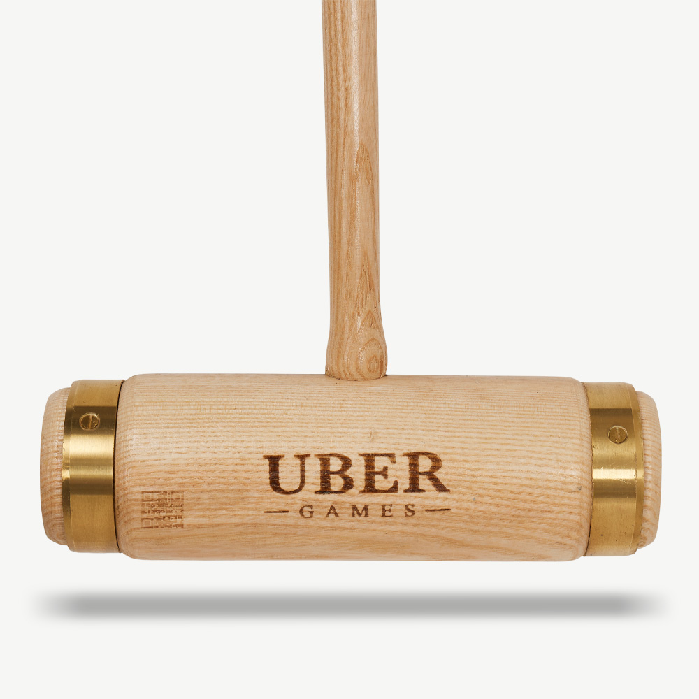 Ein hölzerner Krocket-Schläger. Er hat einen hellbraunen Griff und Kopf mit Messingringen. Auf dem Kopf steht "UBER GAMES".