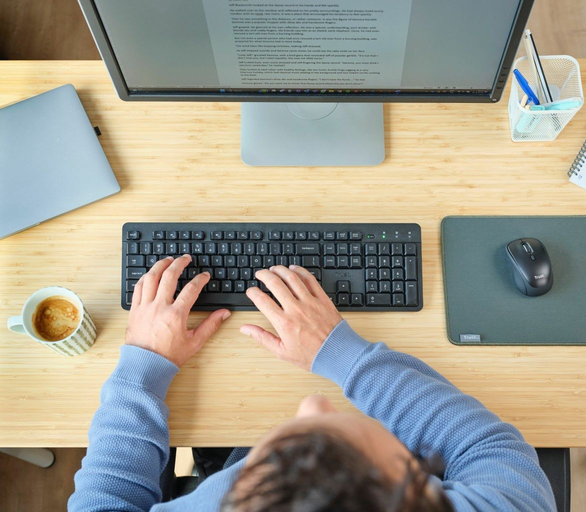Osoba pisząca na klawiaturze, z monitorem, laptopem, filiżanką kawy i myszą. Biurko jest drewniane.
