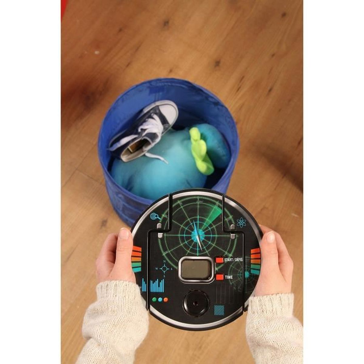 Een persoon houdt een zwart elektronisch apparaat met een scherm vast. Op de achtergrond bevat een blauwe bak een schoen en een knuffel.