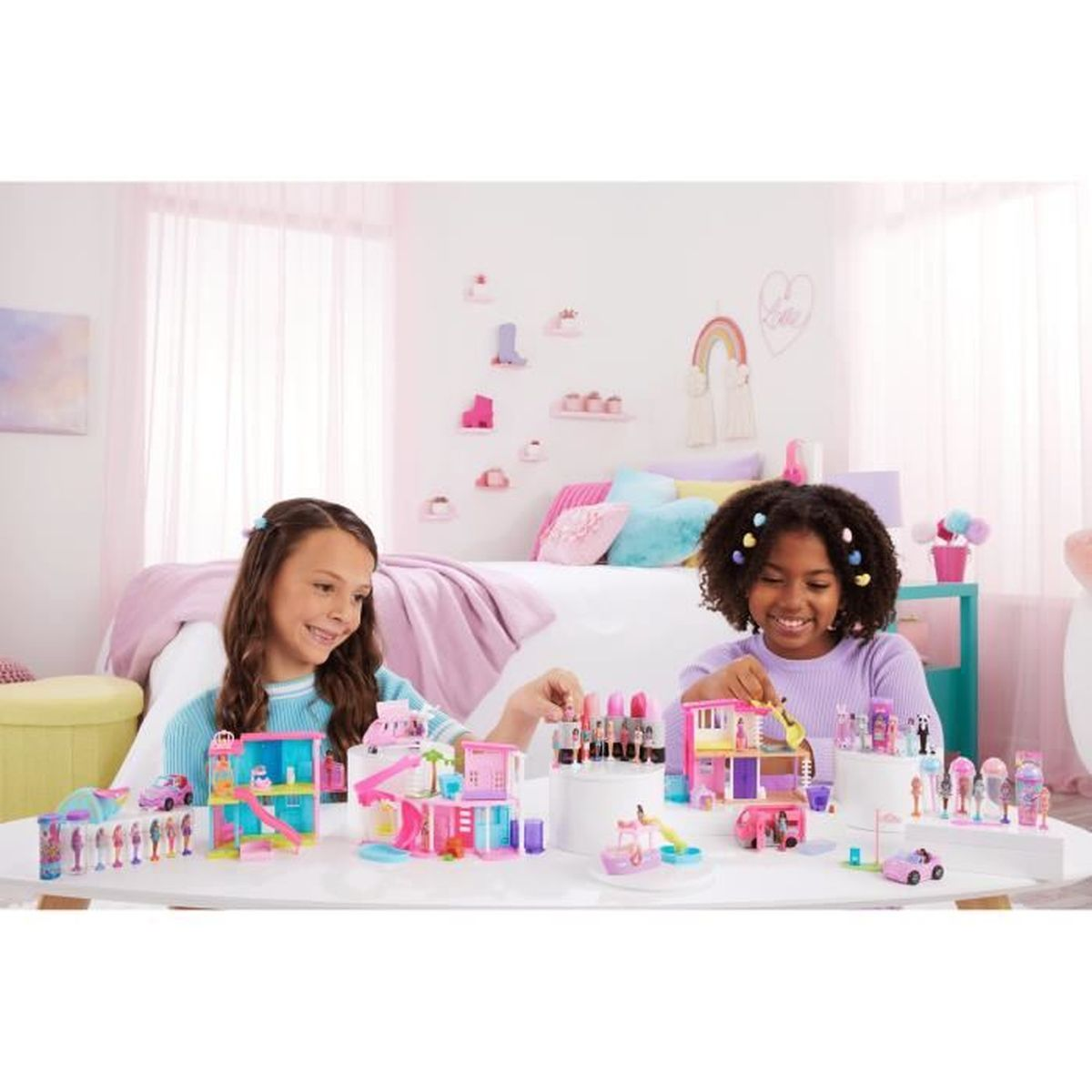 Dos chicas juegan con casas de muñecas y accesorios sobre una mesa blanca. El fondo muestra un dormitorio.