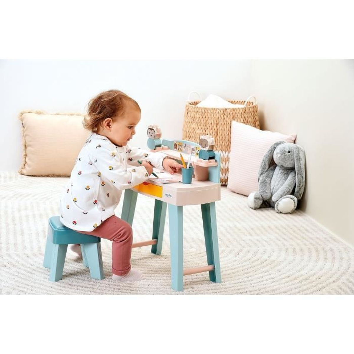 Un bambino è seduto a una scrivania che disegna. La scrivania è azzurra e beige. Un coniglietto di peluche e cuscini sullo sfondo.