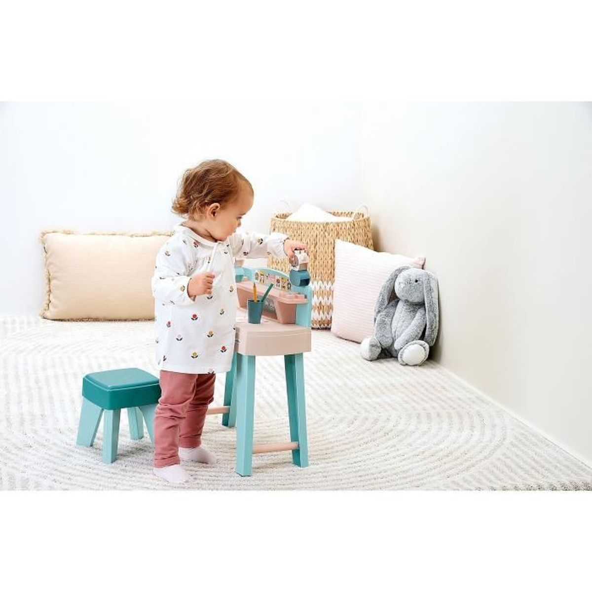 Un bambino piccolo con una camicia bianca con stampa floreale gioca con un banco da lavoro giocattolo. Uno sgabello blu e un coniglietto di peluche grigio sono vicini.