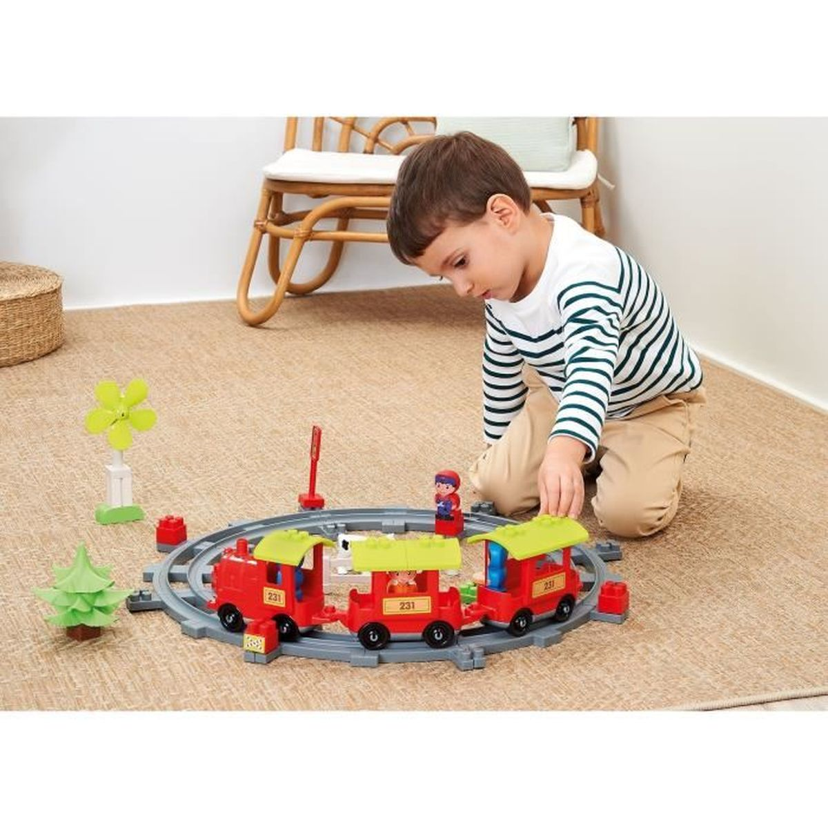 Un bambino gioca con un trenino giocattolo su un tappeto. Il treno è rosso e verde. C'è un albero e un mulino a vento giocattolo nelle vicinanze.