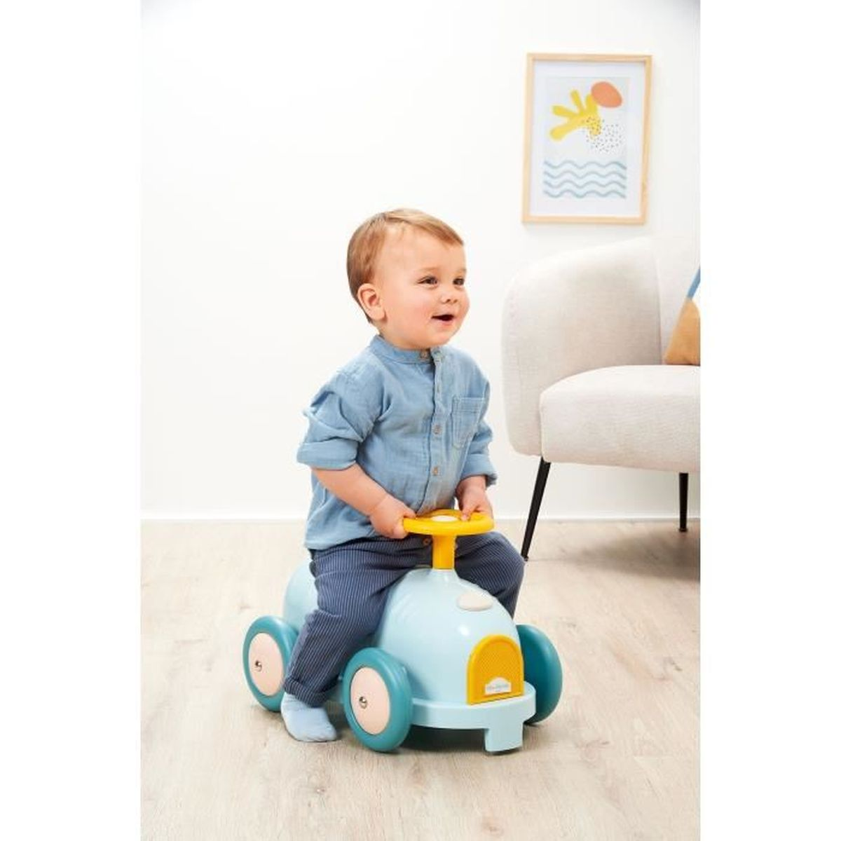 Un bambino piccolo è seduto su un'auto giocattolo blu, sorridendo. Una sedia e una cornice sullo sfondo.