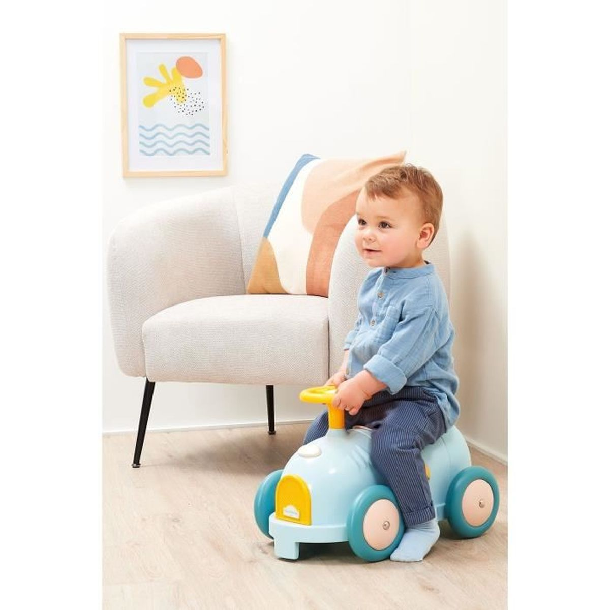 Un bambino seduto su un'auto giocattolo blu con dettagli gialli. Una sedia e un'opera d'arte incorniciata sono sullo sfondo.