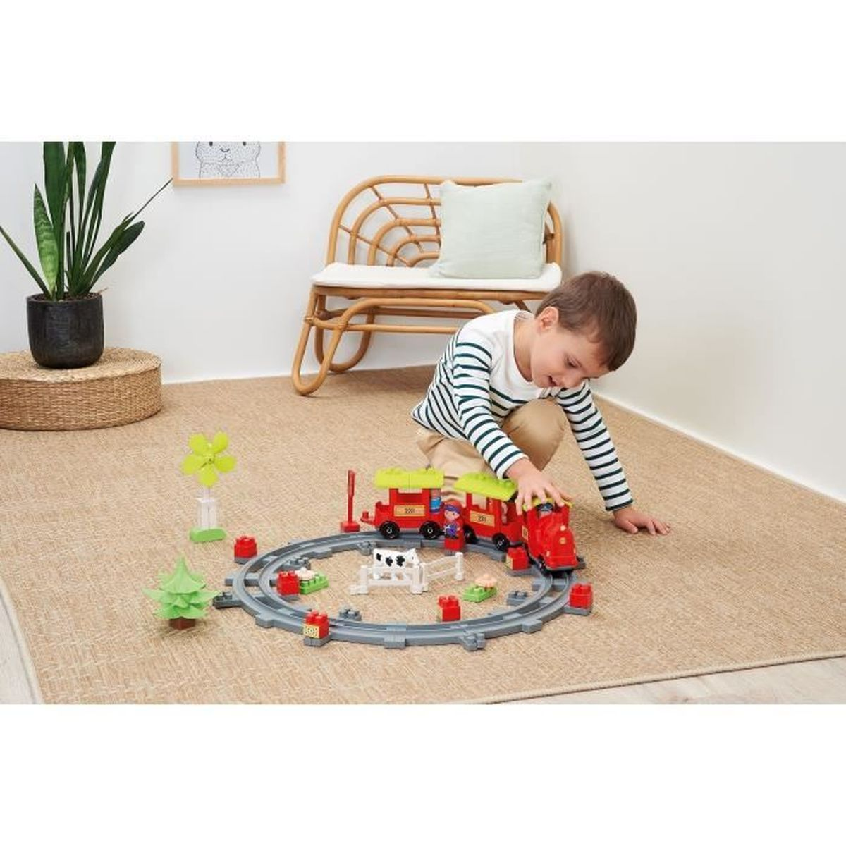 Un bambino gioca con un trenino giocattolo su un tappeto. Il set include un treno rosso, binari e animali della fattoria.