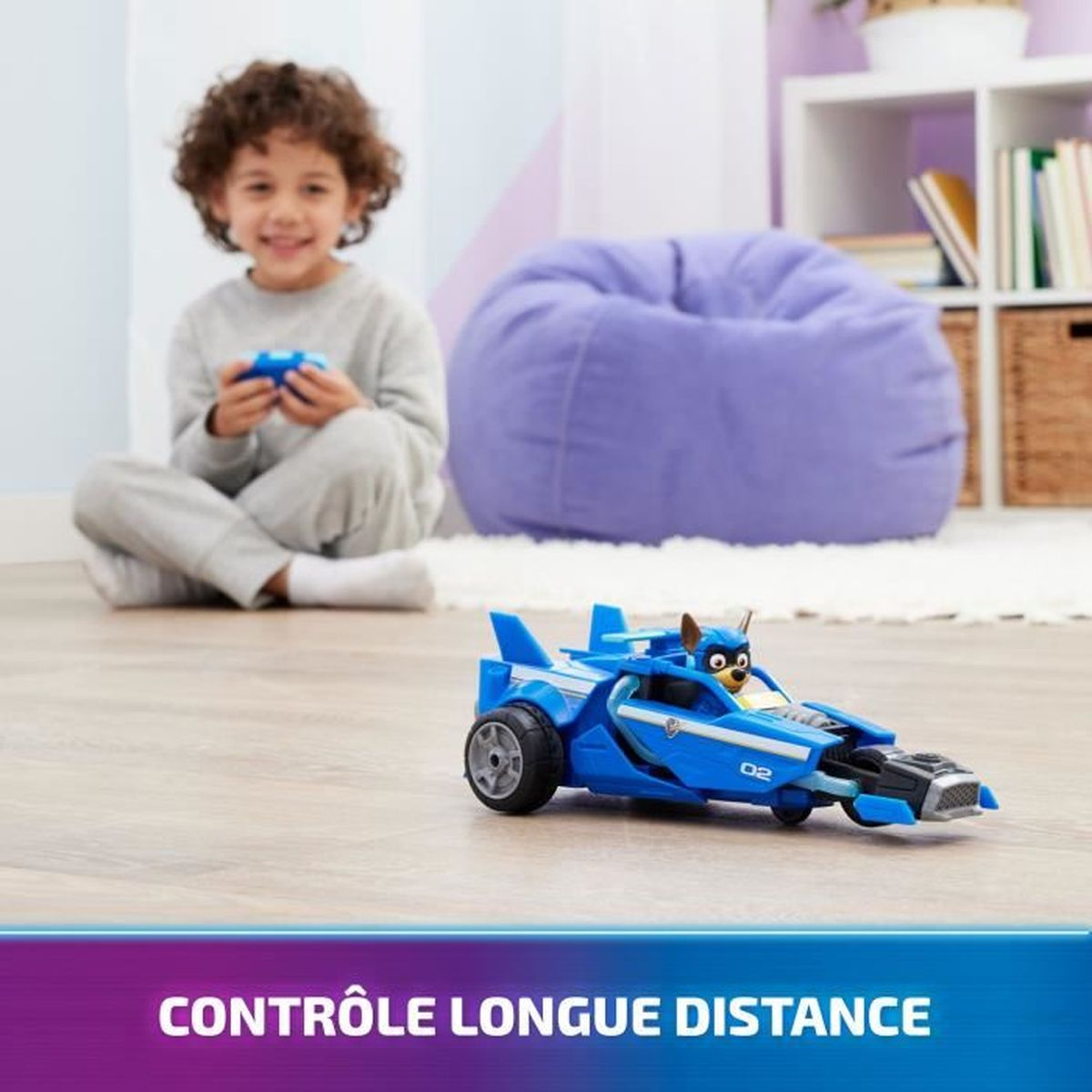 Dziecko uśmiecha się, kontrolując niebieski samochodzik. W tle pufa i półki. Tekst: kontrola na duże odległości.