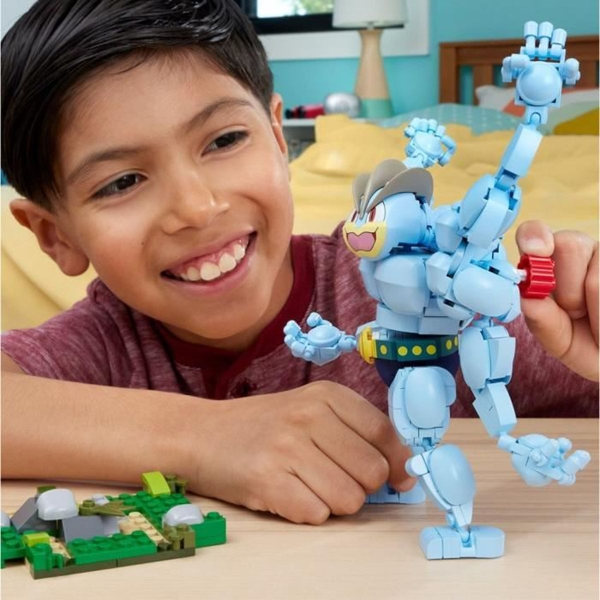Un ragazzo sorride, tenendo in mano un robot giocattolo blu. Un paesaggio Lego è sul tavolo. Lo sfondo è una parete gialla con una lampada.