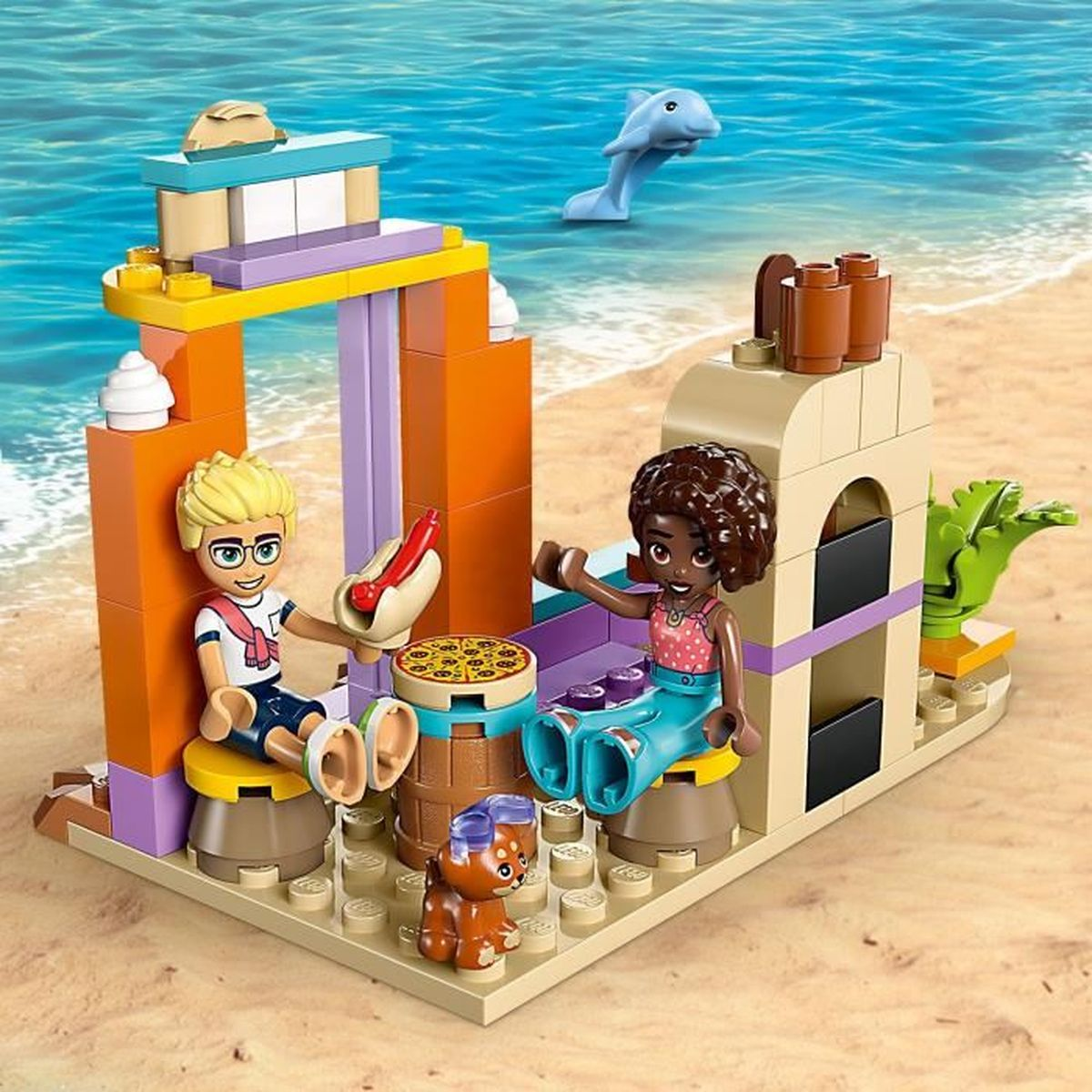 Scena Lego na plaży z dwiema postaciami, delfinem i jedzeniem. Jasne kolory: pomarańczowy, żółty, niebieski. Postacie siedzą.