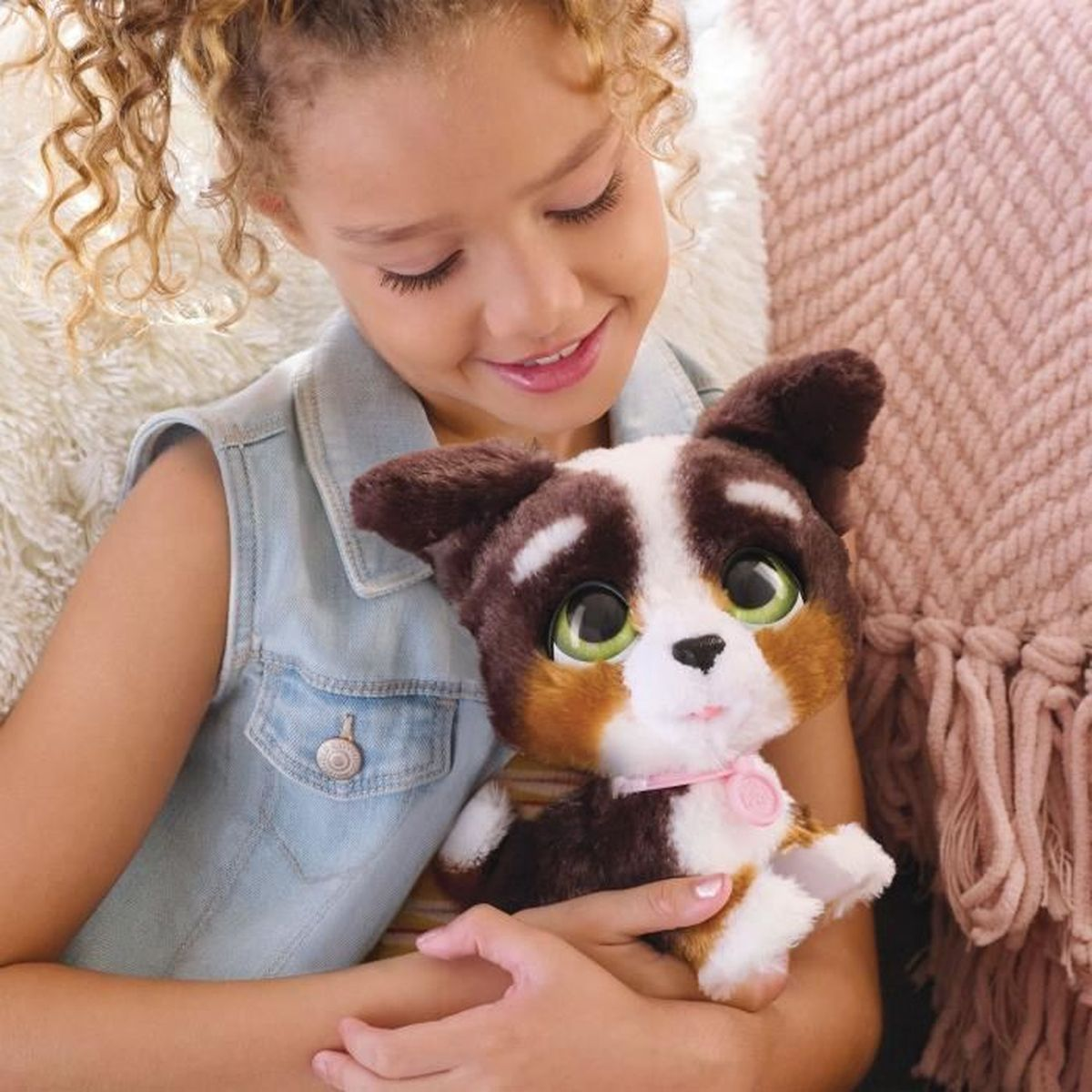 Une fille sourit en tenant un jouet de chiot marron, blanc et beige. La fille porte un gilet en jean. Une couverture rose est en arrière-plan.