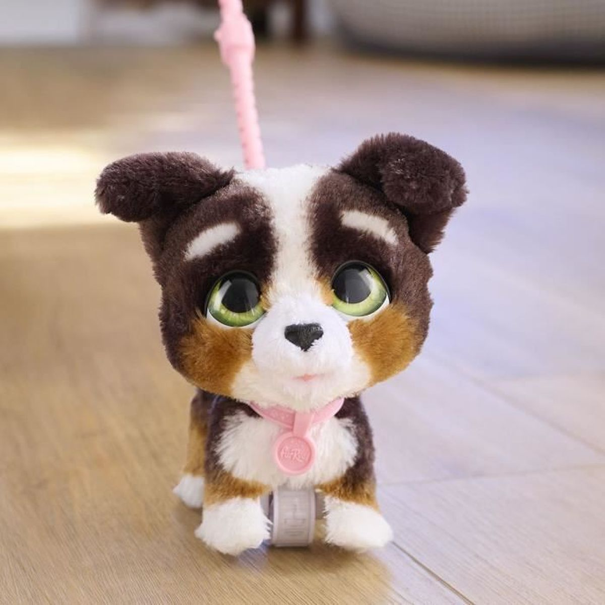 Un jouet en peluche de chiot avec une fourrure marron, blanche et beige. Il a de grands yeux verts et un collier rose. Il est posé sur un sol en bois.