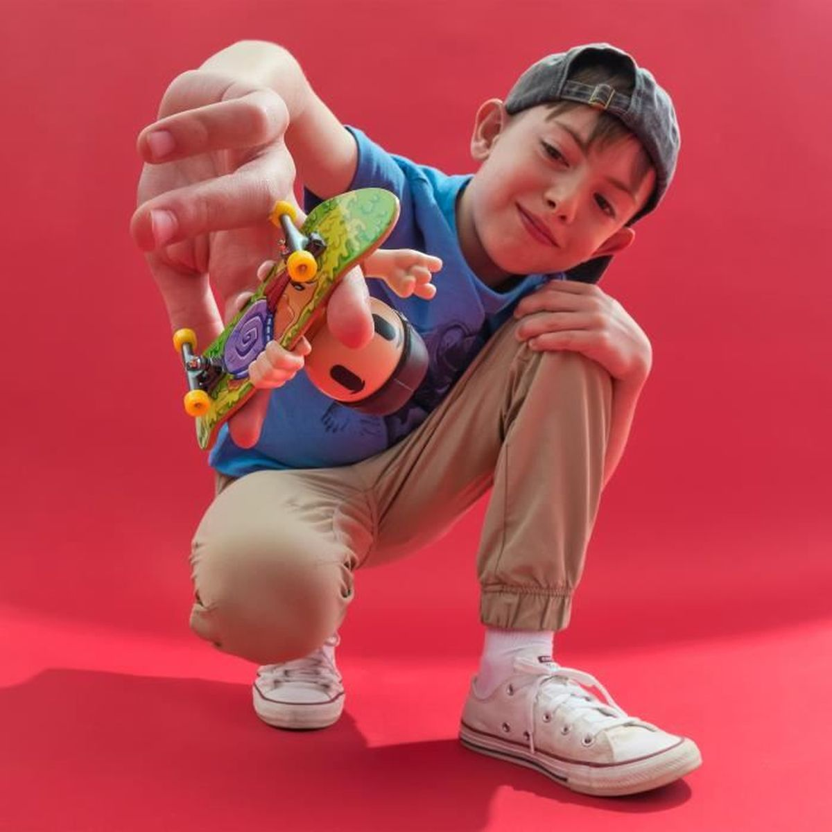 Chłopiec w niebieskiej koszuli i czapce trzyma małą deskorolkę na czerwonym tle, kuca. Ma na sobie białe buty i spodnie khaki.