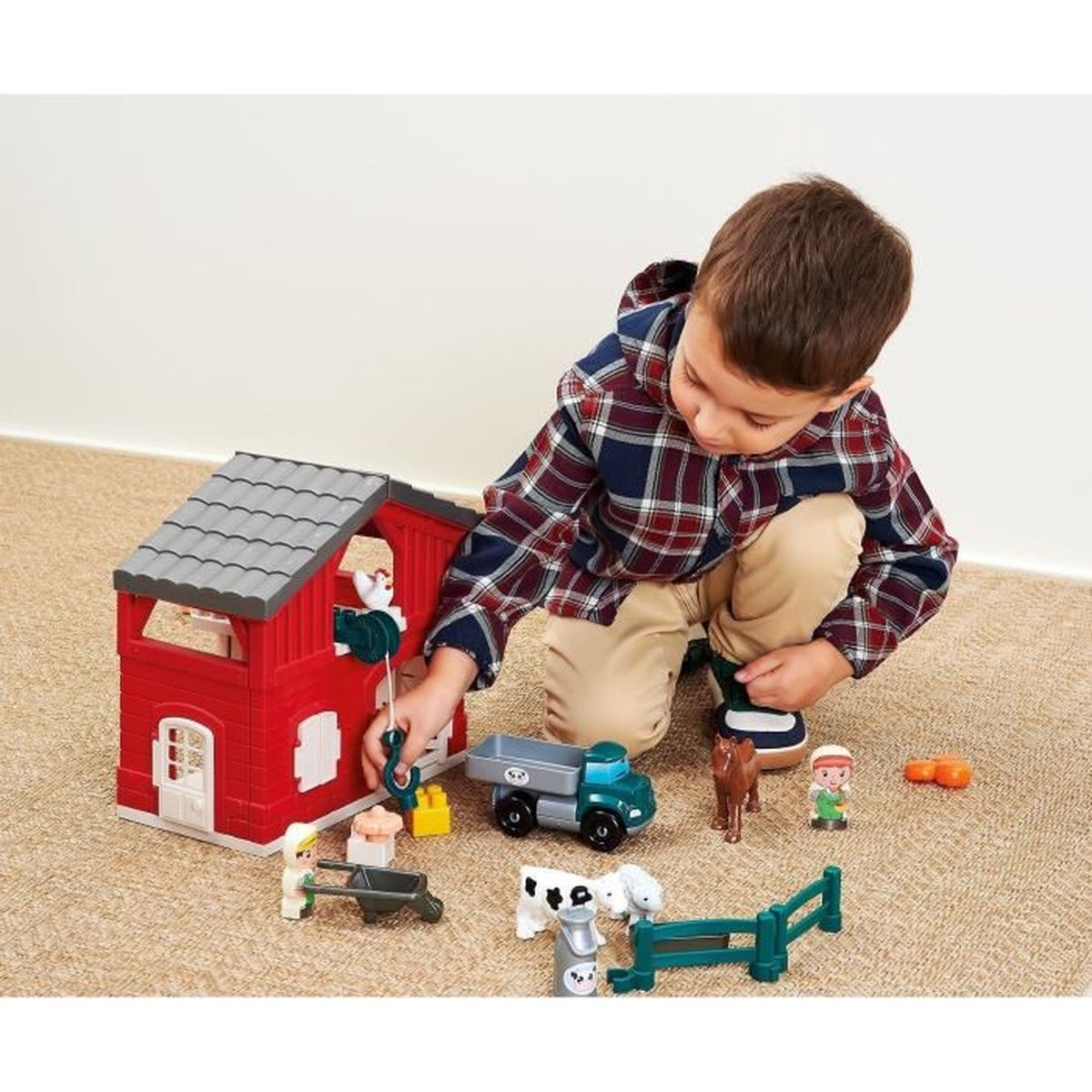 Un bambino gioca con un set di fattoria giocattolo: fienile rosso, camion, animali e figure di contadini su un tappeto beige. Il bambino indossa una camicia a quadri.