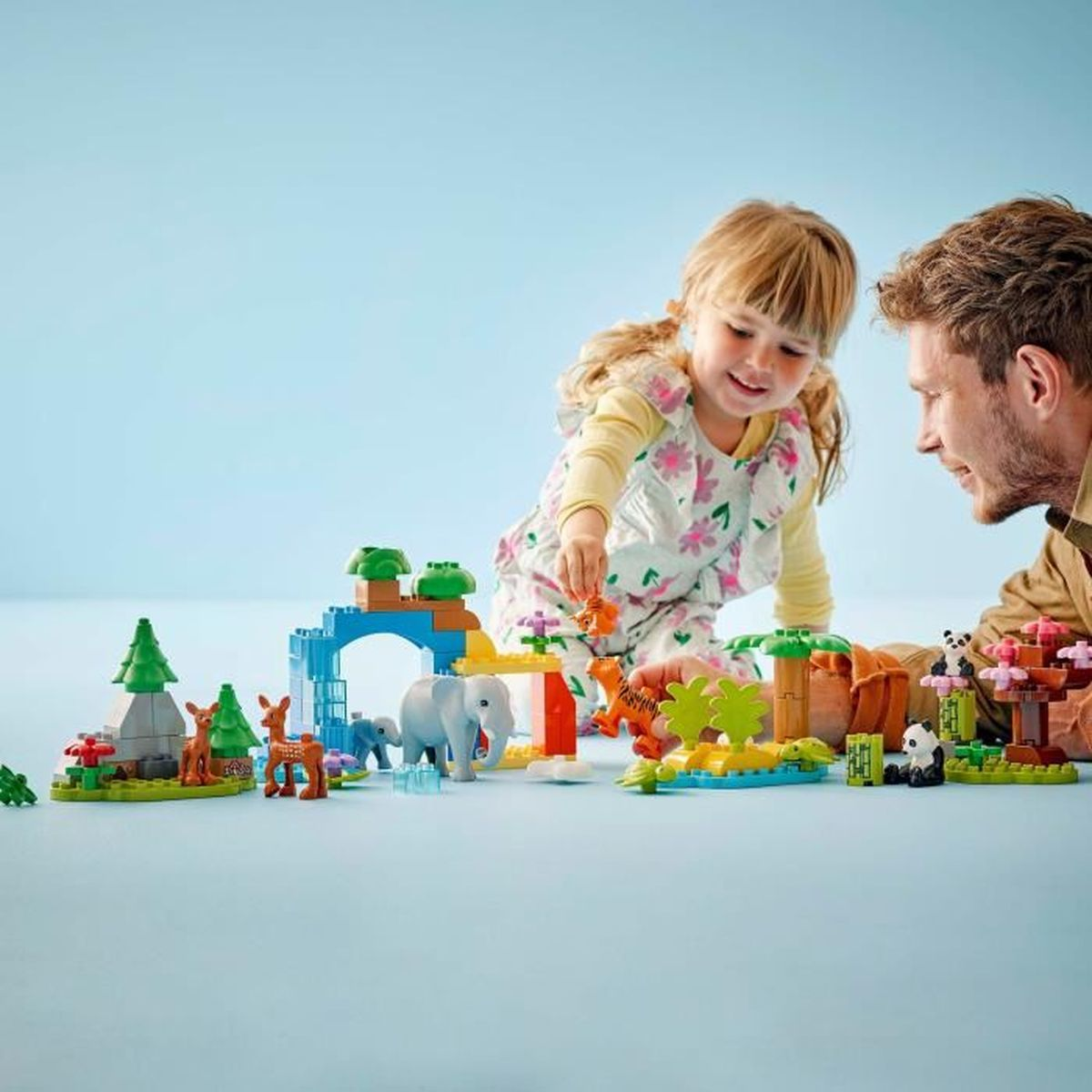 Ein Kind und ein Mann spielen mit bunten Bausteinen und erschaffen eine Zoo-Szene mit Tieren wie Tiger, Elefant und Panda.