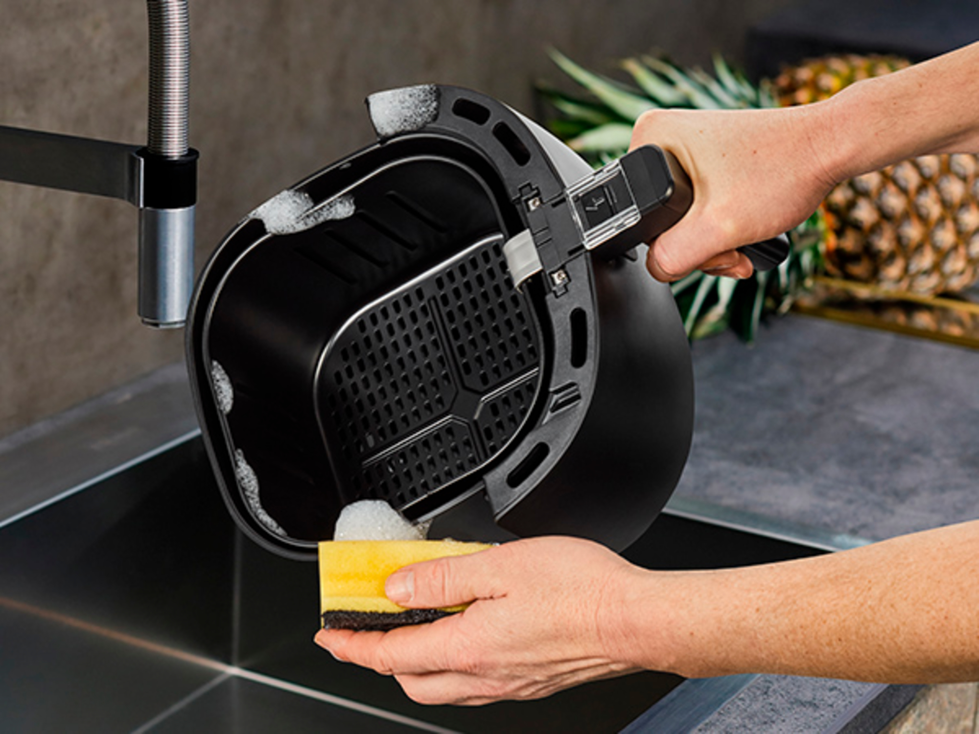 Ręce myją czarną frytkownicę powietrzną żółtą gąbką i mydłem. Ananas i zlewozmywak ze stali nierdzewnej w tle.