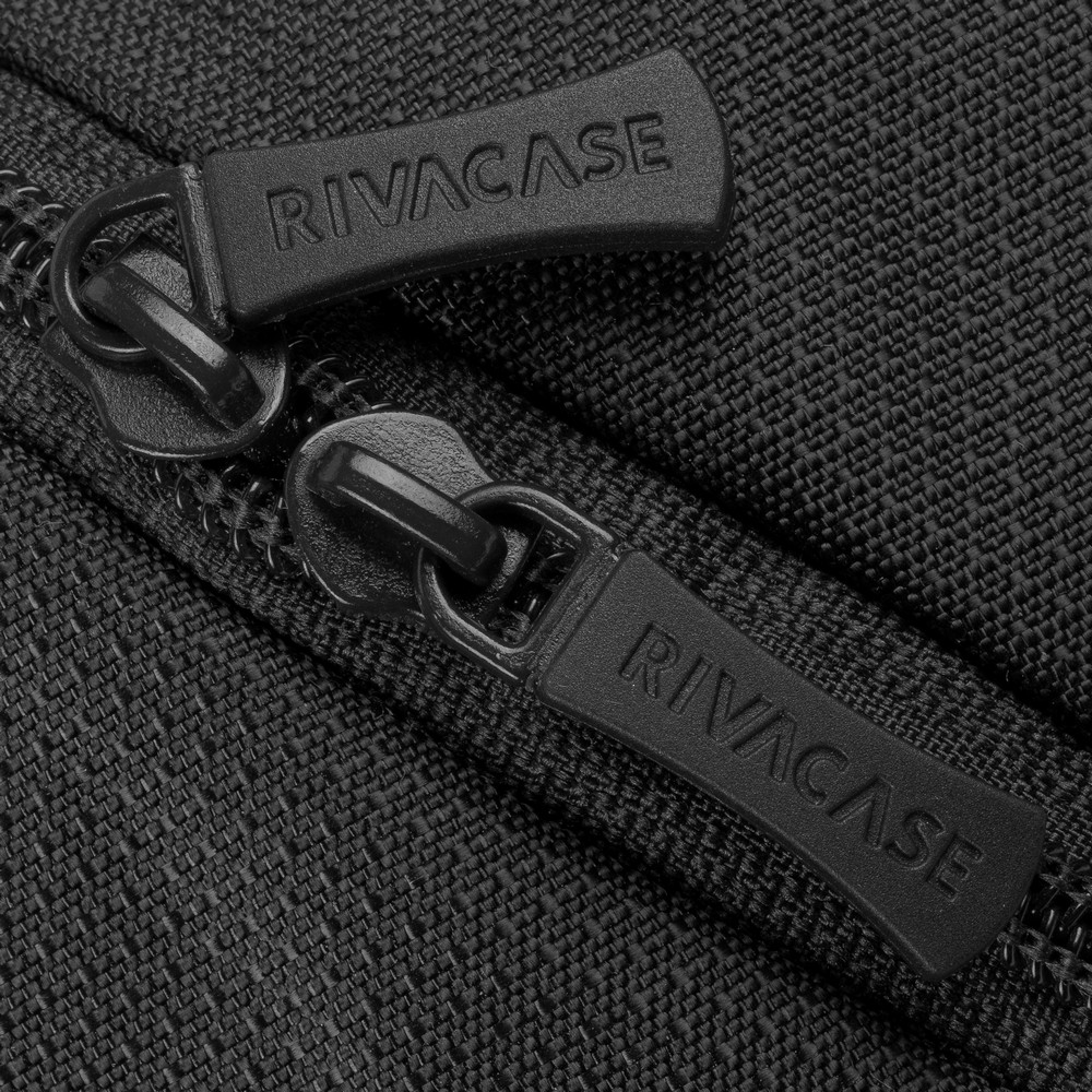 Primo piano di una cerniera nera con 'RIVACASE' stampato sulle linguette. Il tessuto è un materiale grigio scuro strutturato.