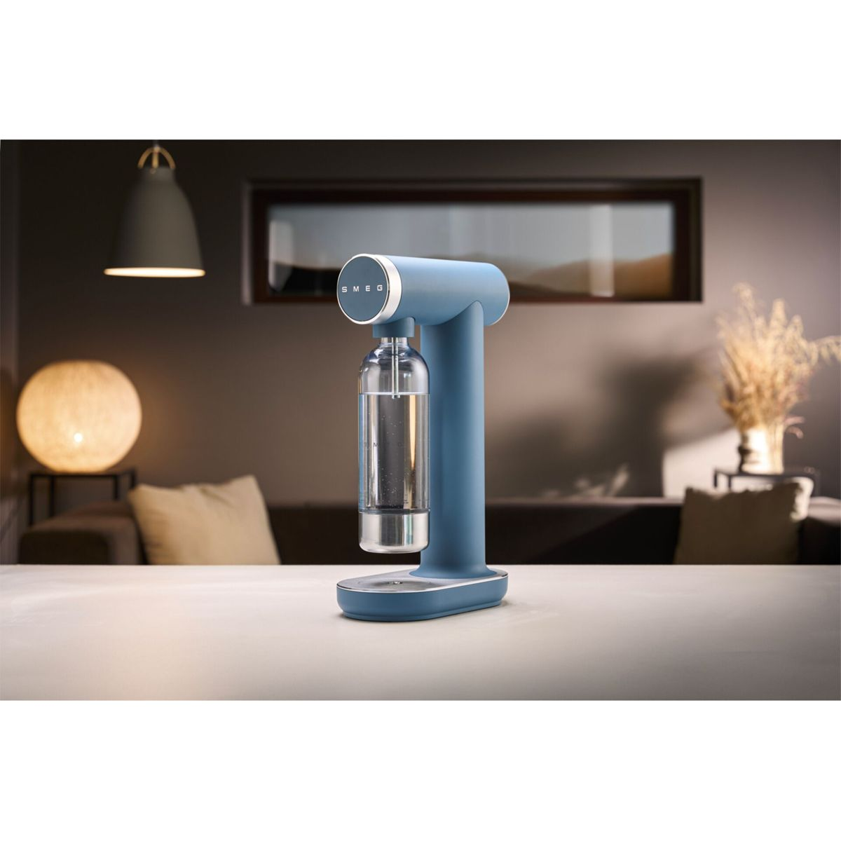 Ein blauer SMEG-Soda-Maker auf einem weißen Tisch. Eine Glasflasche ist befestigt. Im Hintergrund ist ein Wohnzimmer mit Lampe.