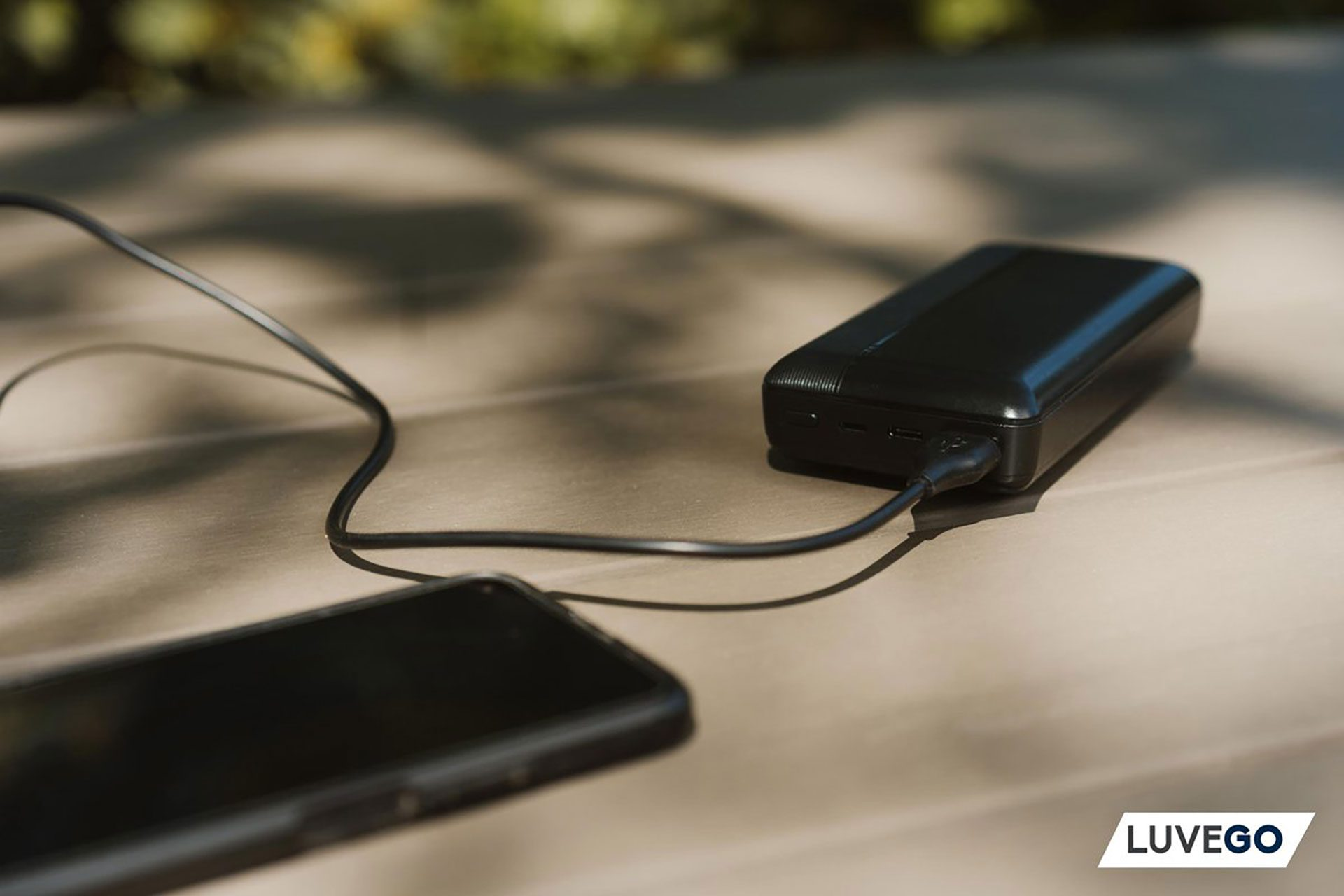 Eine schwarze Powerbank ist mit einem Ladekabel mit einem Telefon verbunden. Telefon und Powerbank liegen auf einer Holzoberfläche.