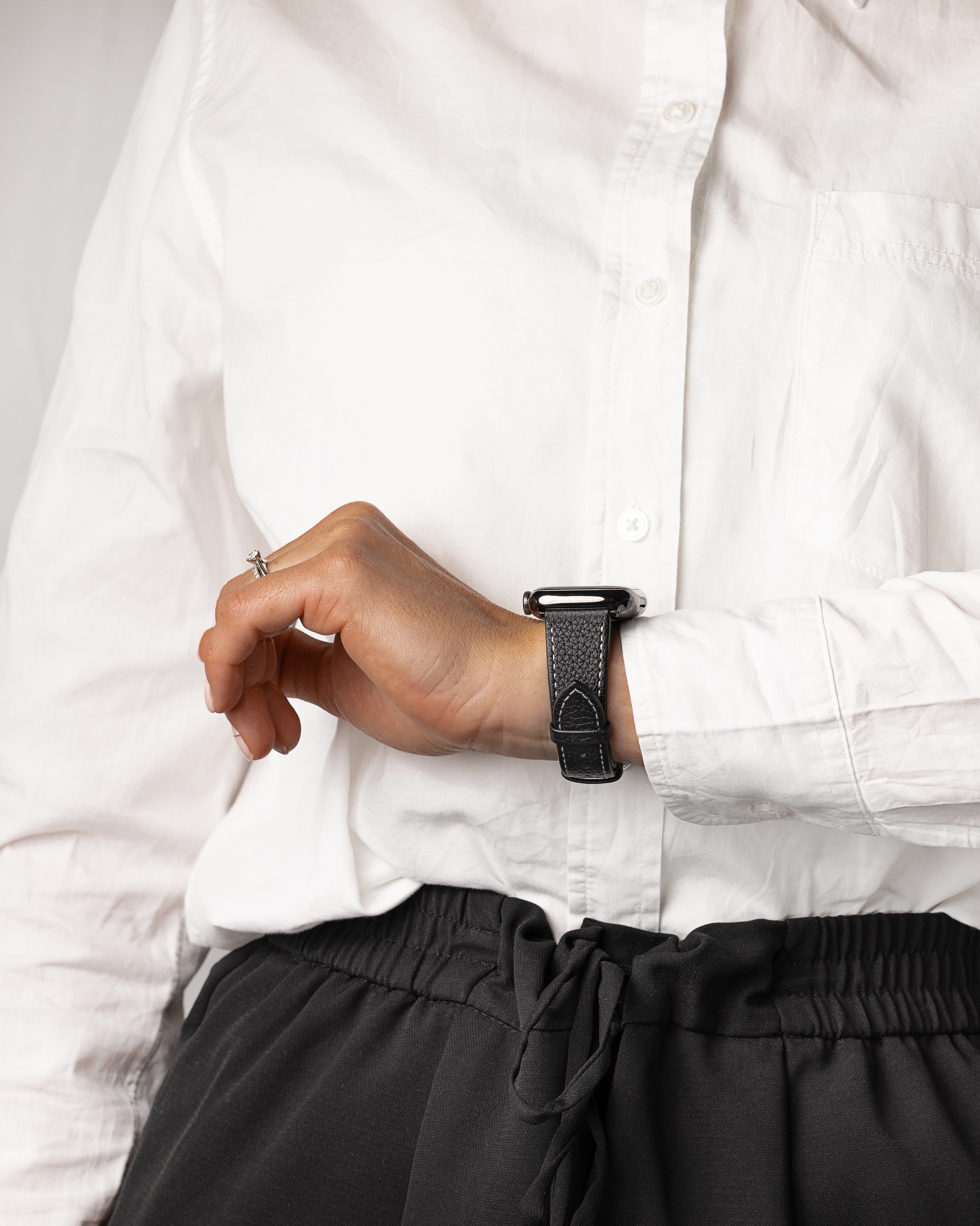 Eine Person mit weißem Hemd und schwarzer Hose trägt eine schwarze Uhr mit schwarzem Armband. Ein Ring ist an ihrer Hand.