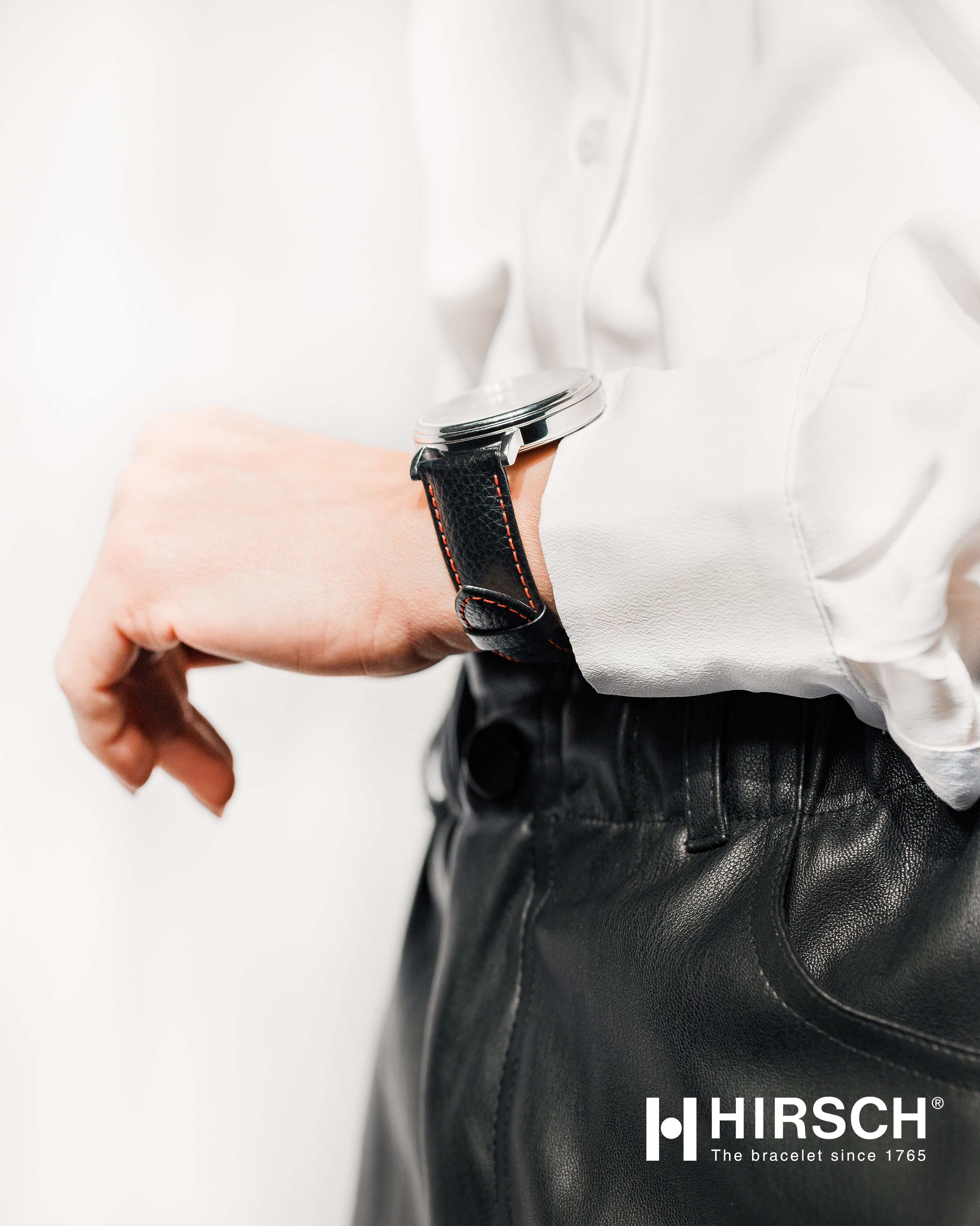 Eine Person, die eine Uhr mit schwarzem Lederarmband und roten Nähten trägt. Die Person trägt ein weißes Hemd und eine schwarze Hose.