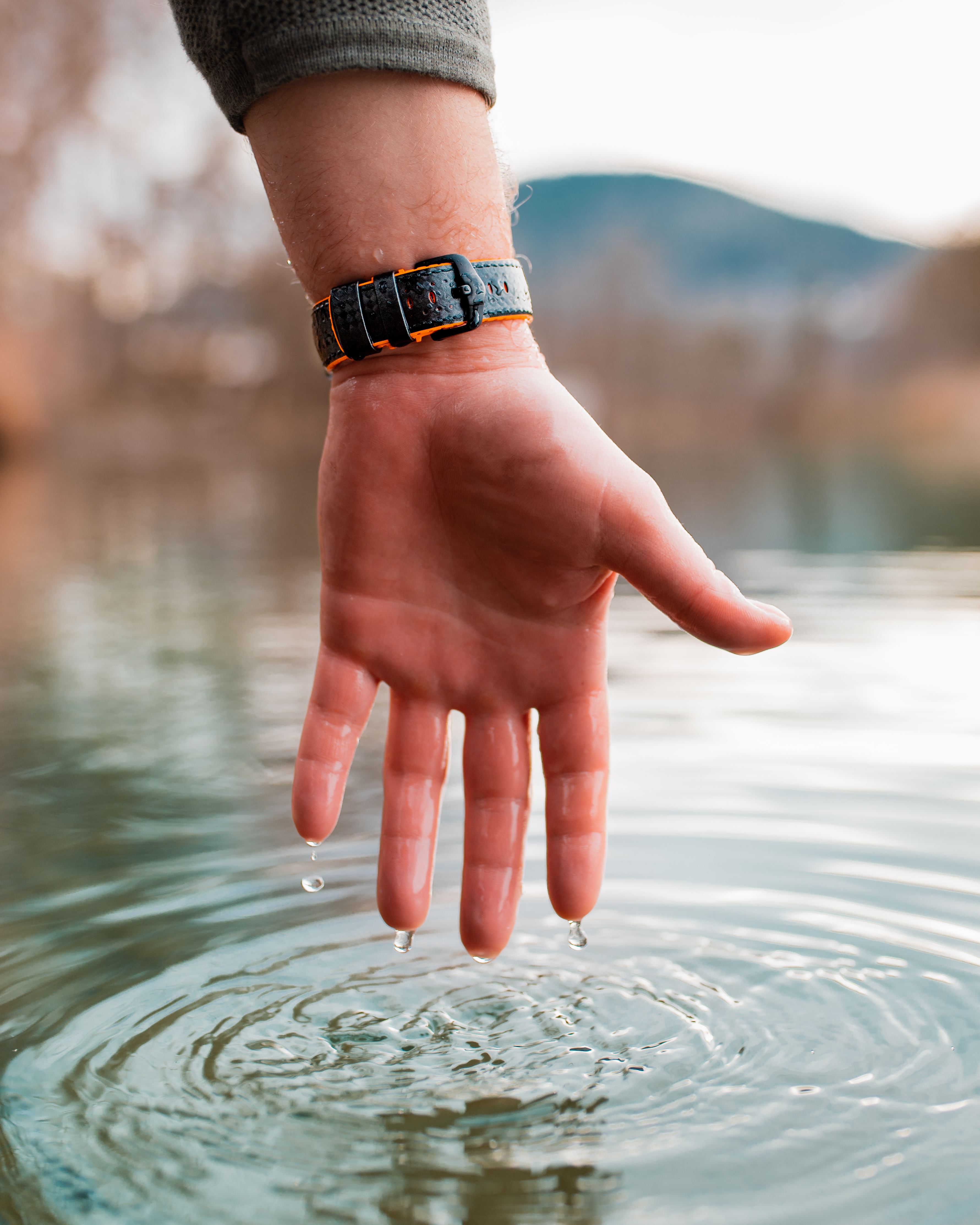 Hand, die sich dem Wasser nähert, Tropfen fallen. Am Handgelenk ist eine Uhr. Wellen im Wasser. Der Hintergrund ist unscharf.