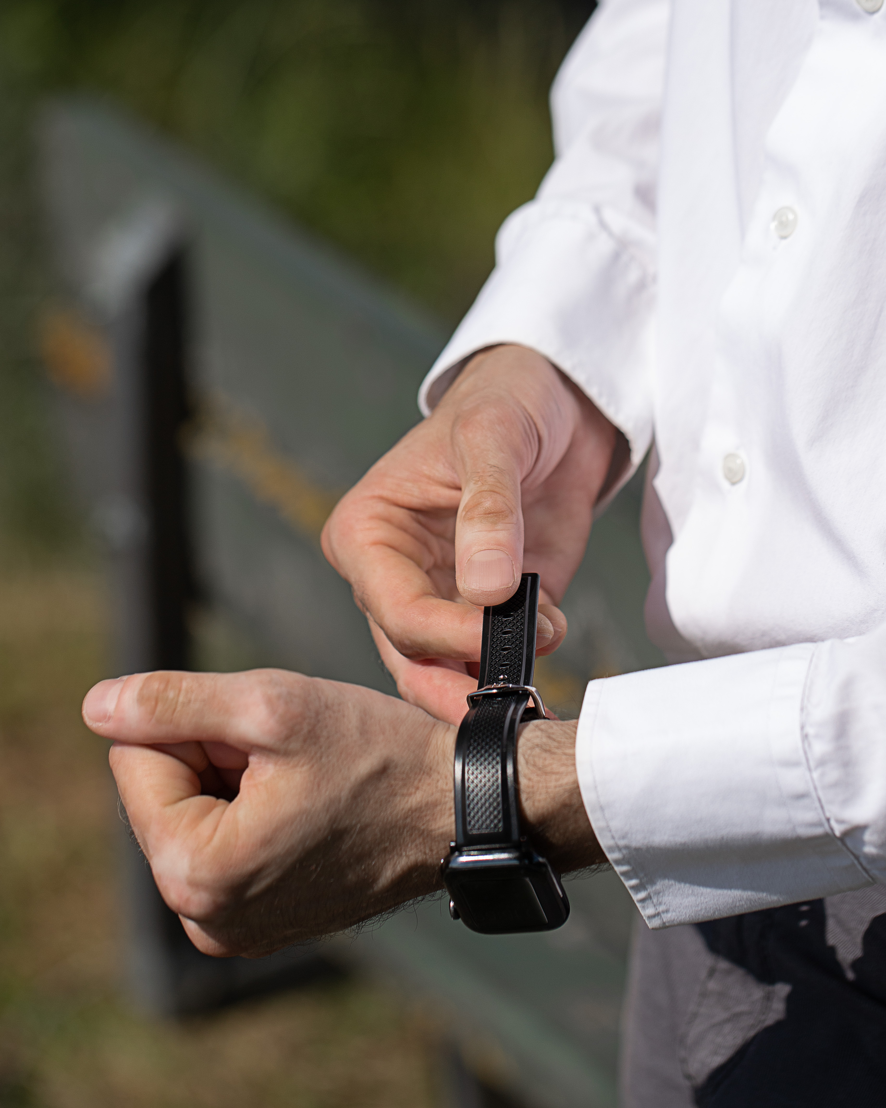 Mann, der eine schwarze Uhr an seinem Handgelenk einstellt. Er trägt ein weißes Hemd. Der Hintergrund ist grün und grau verschwommen.