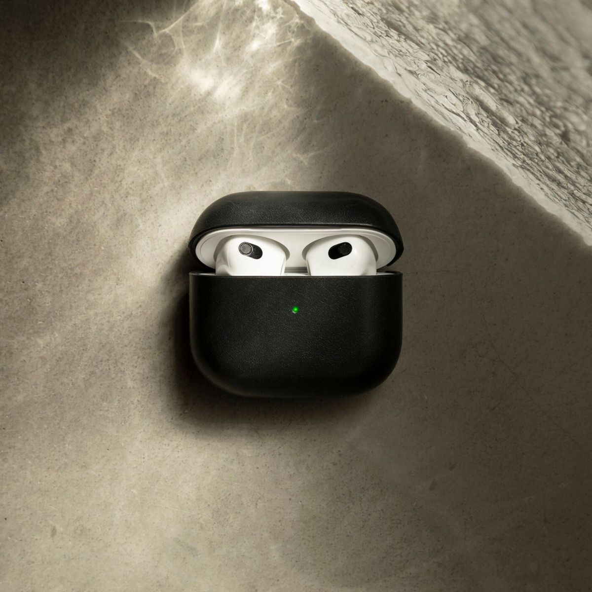 Étui AirPods noir avec écouteurs blancs à l'intérieur, ouvert. Lumière verte sur l'étui. Le fond est une surface gris clair texturée.