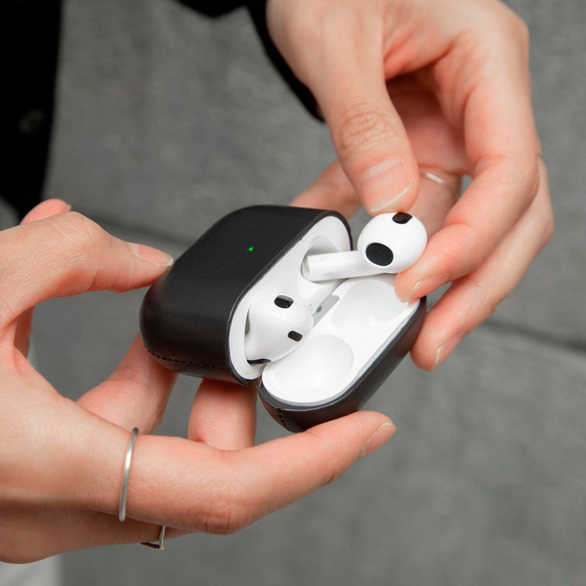 Mains tenant un étui AirPods noir, ouvert, révélant des AirPods blancs. Un AirPod est tenu. Une bague argentée est sur un doigt.