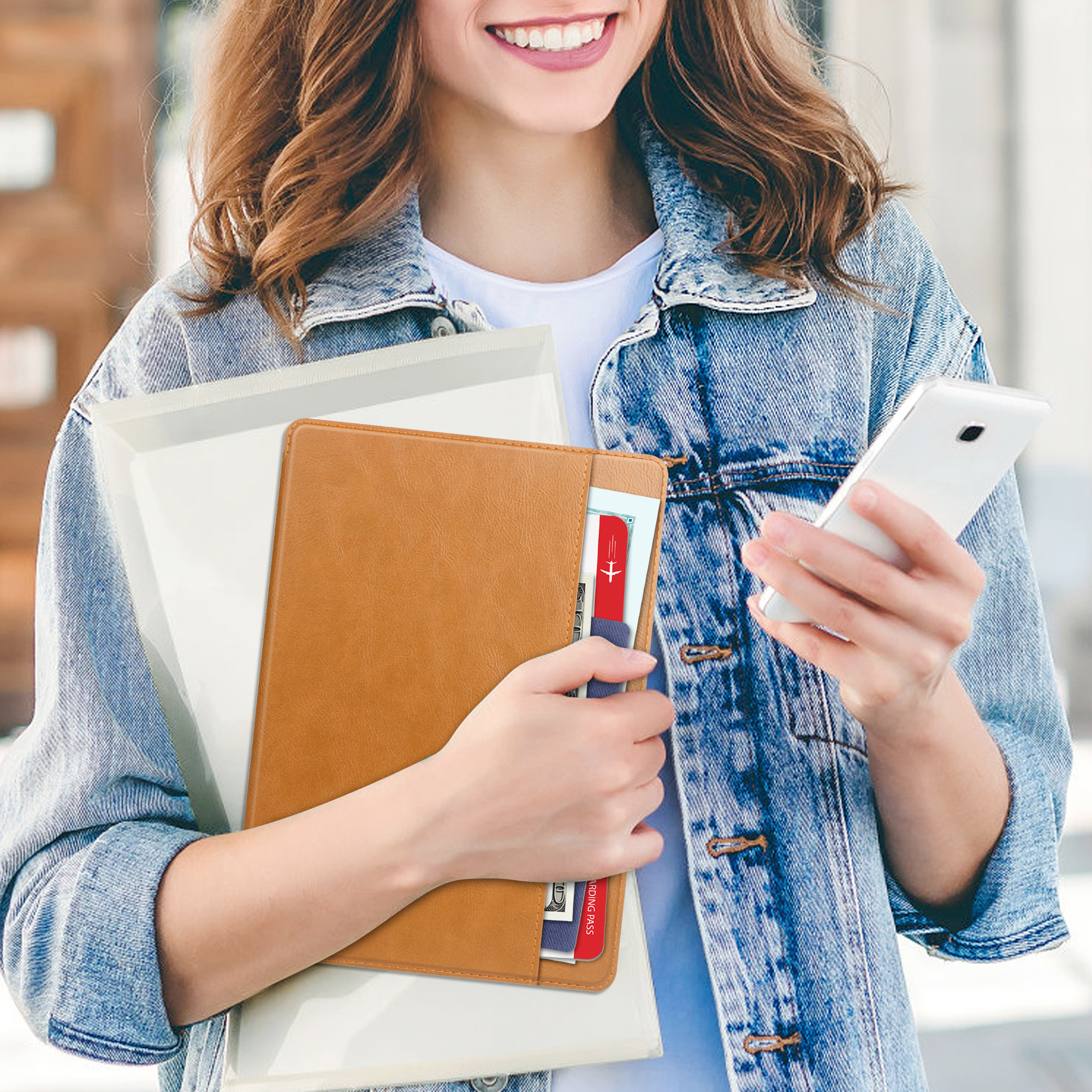 Frau mit Telefon und Dokumenten. Trägt eine Jeansjacke. Lächelt. Hält eine hellbraune Ledermappe.