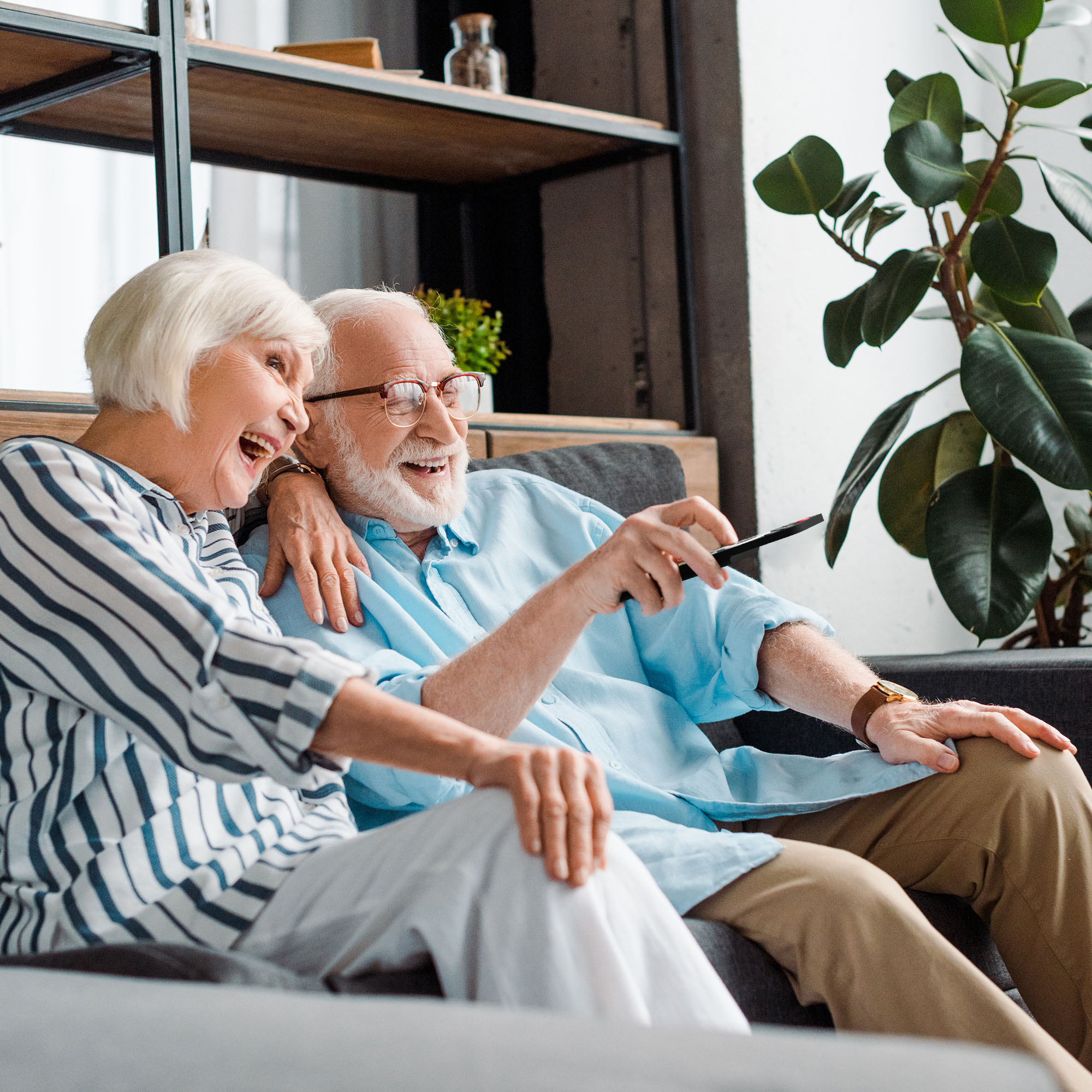 An elderly couple laughs while watching TV. The man holds a remote. They are sitting on a grey couch. A plant is in the background.