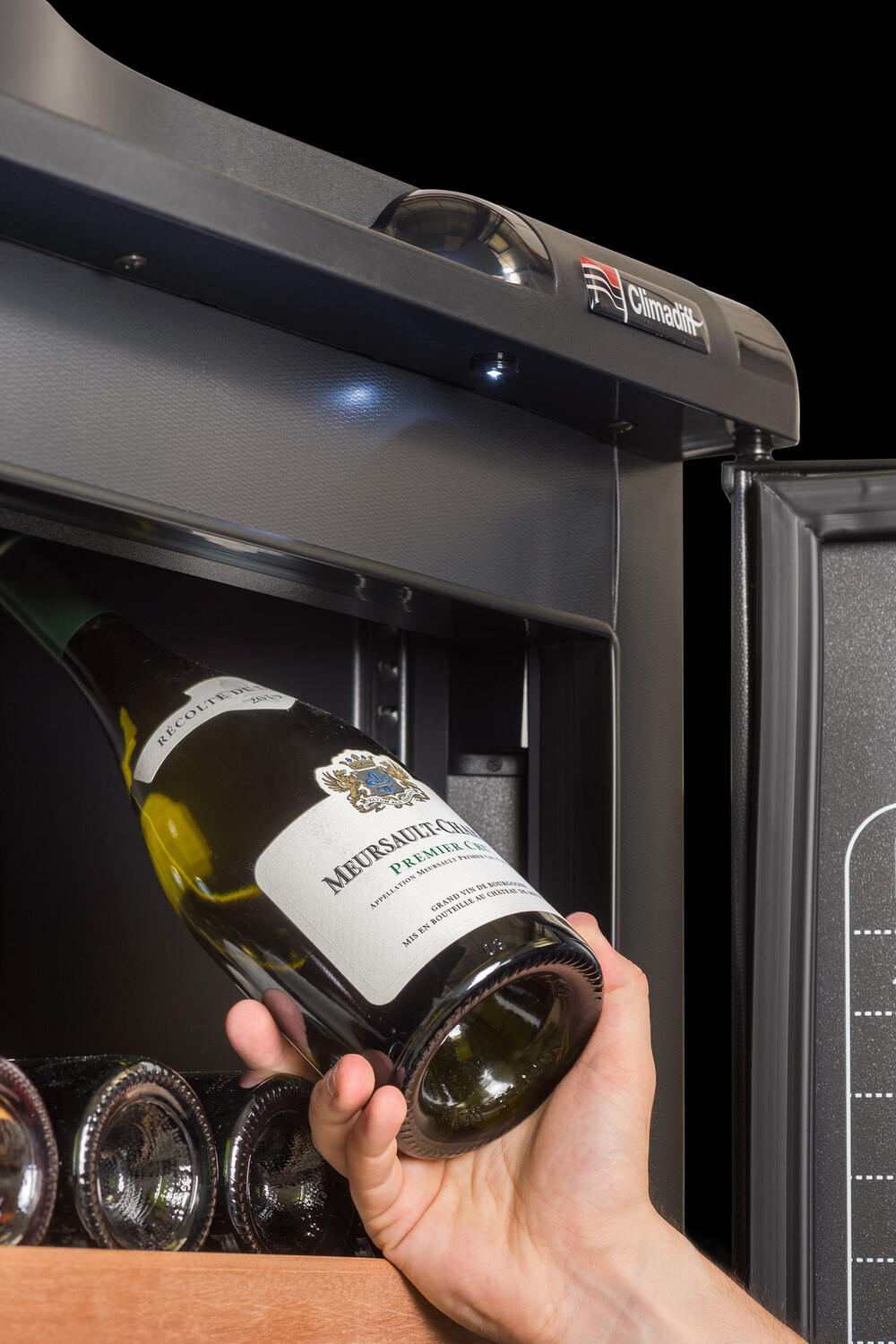 Eine Hand legt eine Weinflasche in einen Weinkühlschrank. Die Flasche hat ein weißes Etikett und steht mit anderen Flaschen auf Holzregalen.
