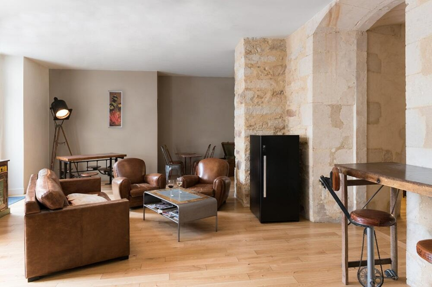 Wohnzimmer mit Ledersofas, einem Couchtisch und einer Scheinwerferlampe. Ein schwarzer Kühlschrank steht an einer Steinmauer.