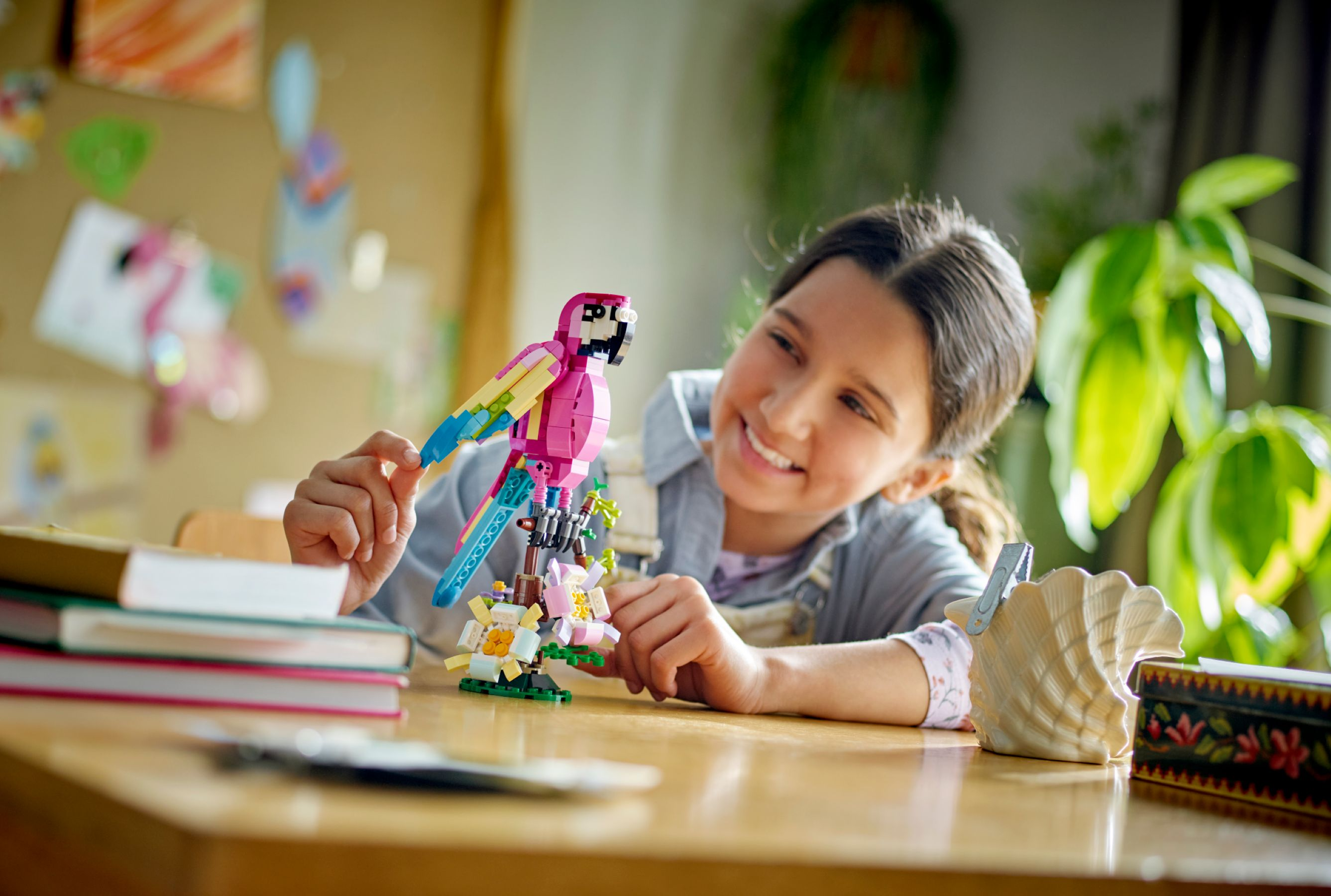 Ein Mädchen lächelt einen Lego-Papagei auf einem Tisch an. Bücher und eine Muschel sind in der Nähe. Hintergrund: Pflanzen, Zeichnungen.