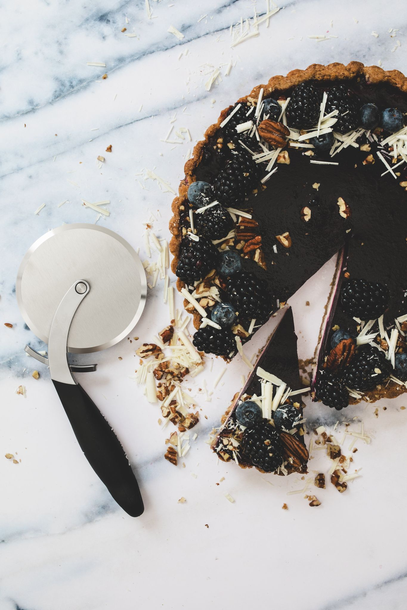 Ein dunkler Beerenkuchen mit einem angeschnittenen Stück, belegt mit Beeren, Nüssen und weißen Schokoladenraspeln, neben einem Pizzaschneider.