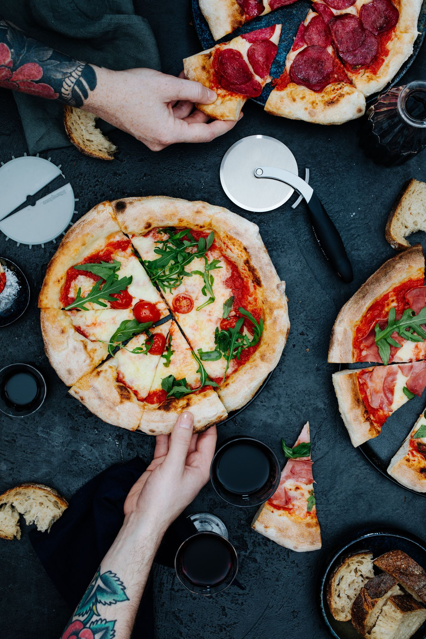 Draufsicht auf Pizzen mit Händen, die nach Stücken greifen. Enthält eine Margherita, Peperoni und Prosciutto Pizza, Brot und Wein.
