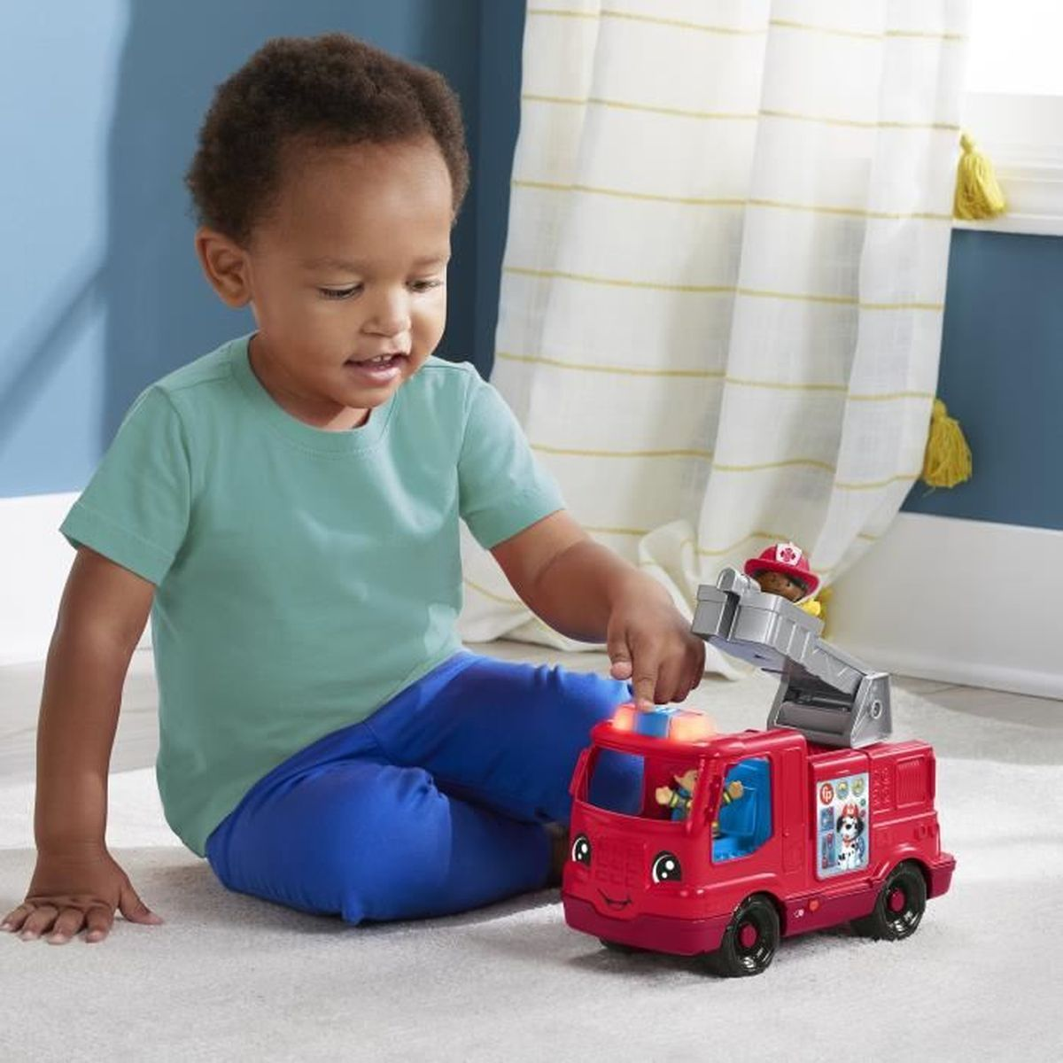 Ein Kind in blauer Hose und grünem Hemd spielt mit einem roten Spielzeug-Feuerwehrwagen. Der Wagen hat eine Leiter und einen Feuerwehrmann.