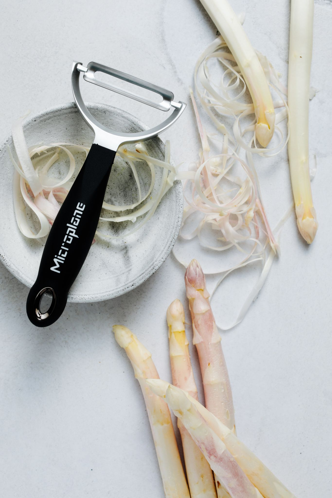 Spargel, Schäler und Späne auf weißer Oberfläche. Einige Spargel sind geschält. Ein schwarzer Schäler und eine kleine Schüssel sind sichtbar.