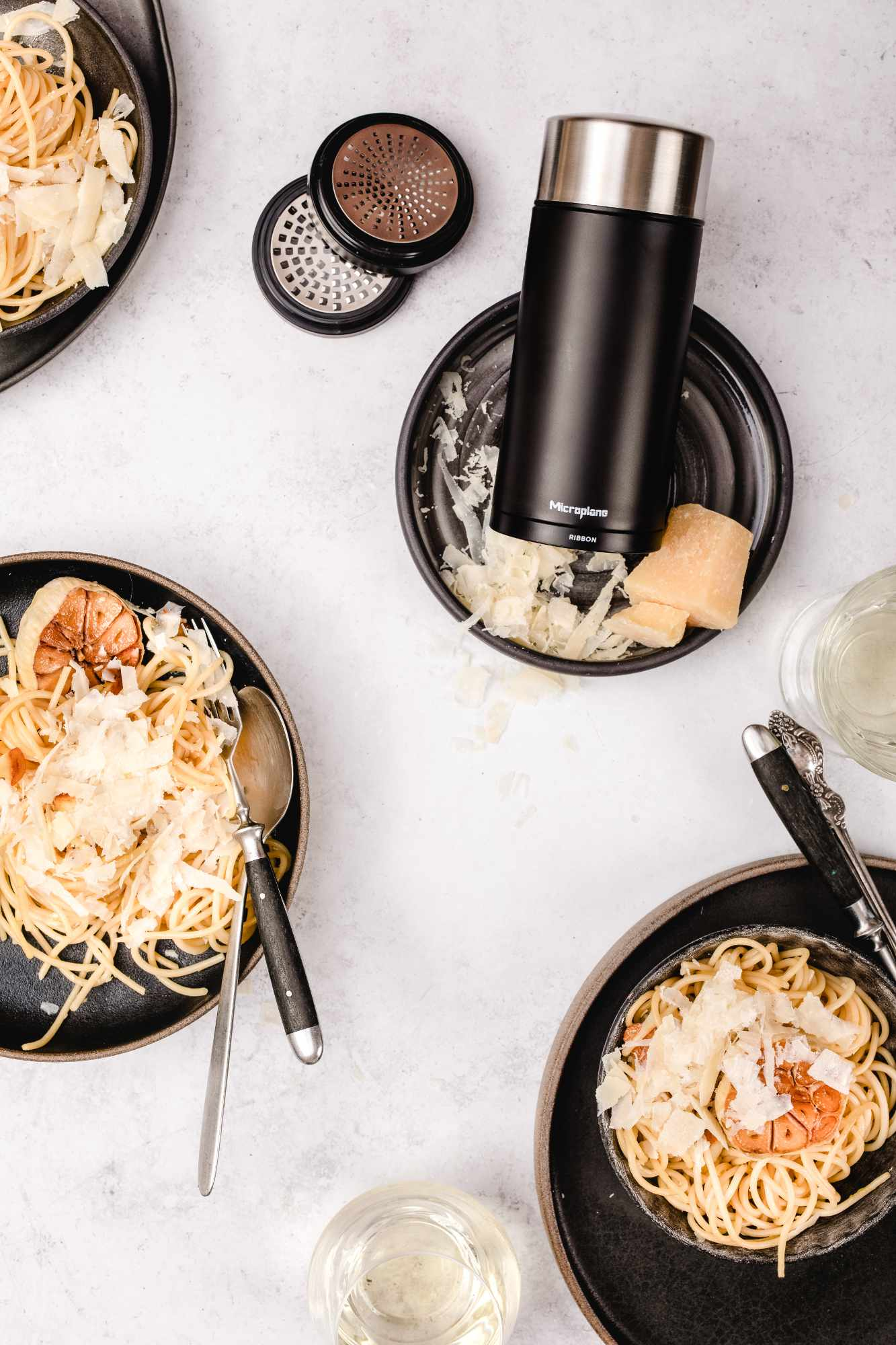 Draufsicht auf Pasta mit Käse, Knoblauch und einem Käsehobel. Schwarze Teller auf weißem Untergrund. Weingläser sind ebenfalls zu sehen.