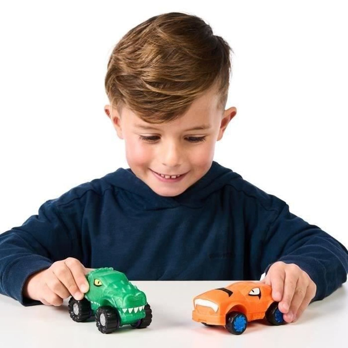 Ein lächelnder Junge hält grüne Alligator- und orangefarbene Tiger-Spielzeugautos. Der Hintergrund ist weiß.